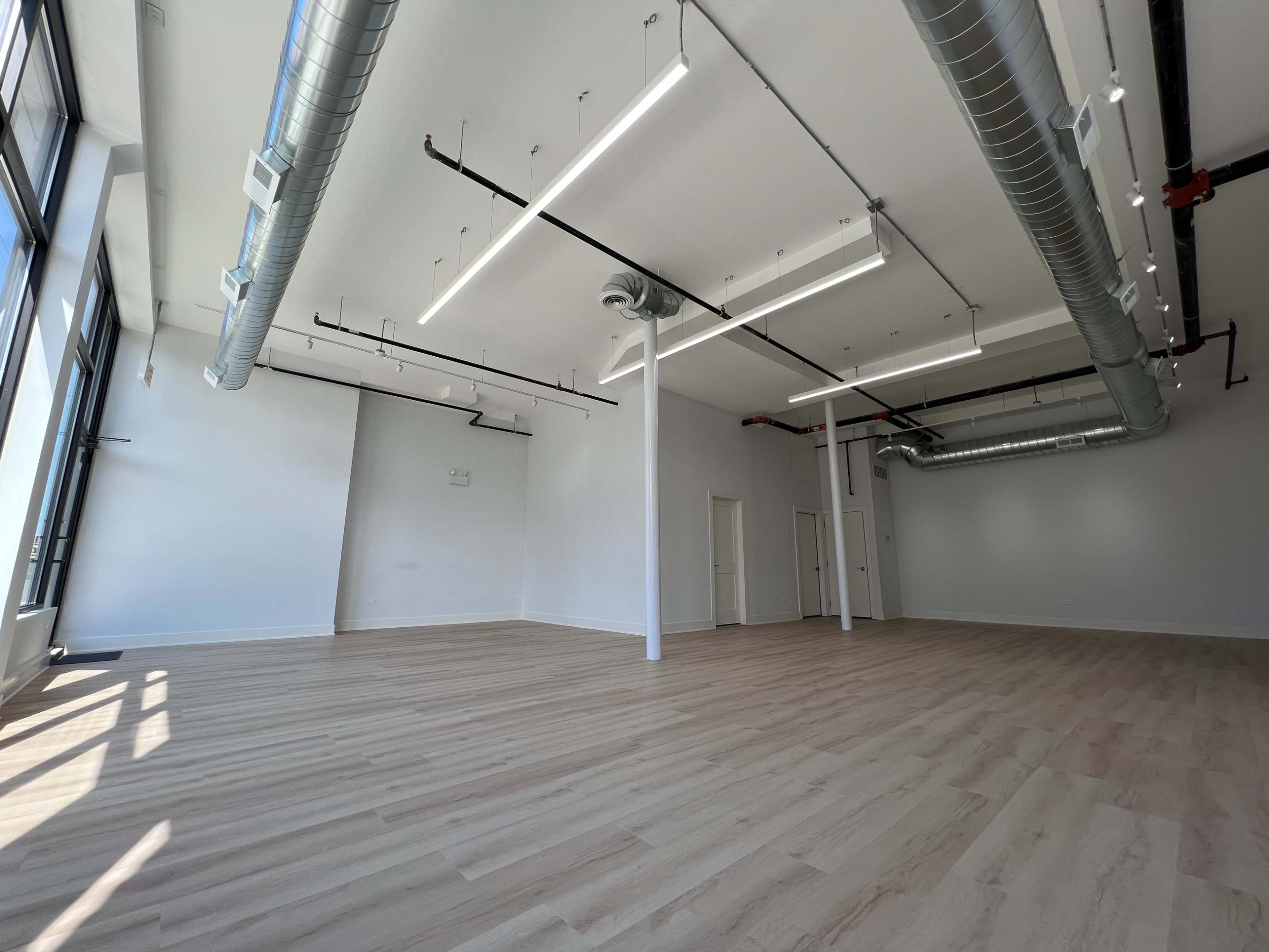 A spacious, empty room with large windows, polished wooden floors, and exposed ductwork on the ceiling.