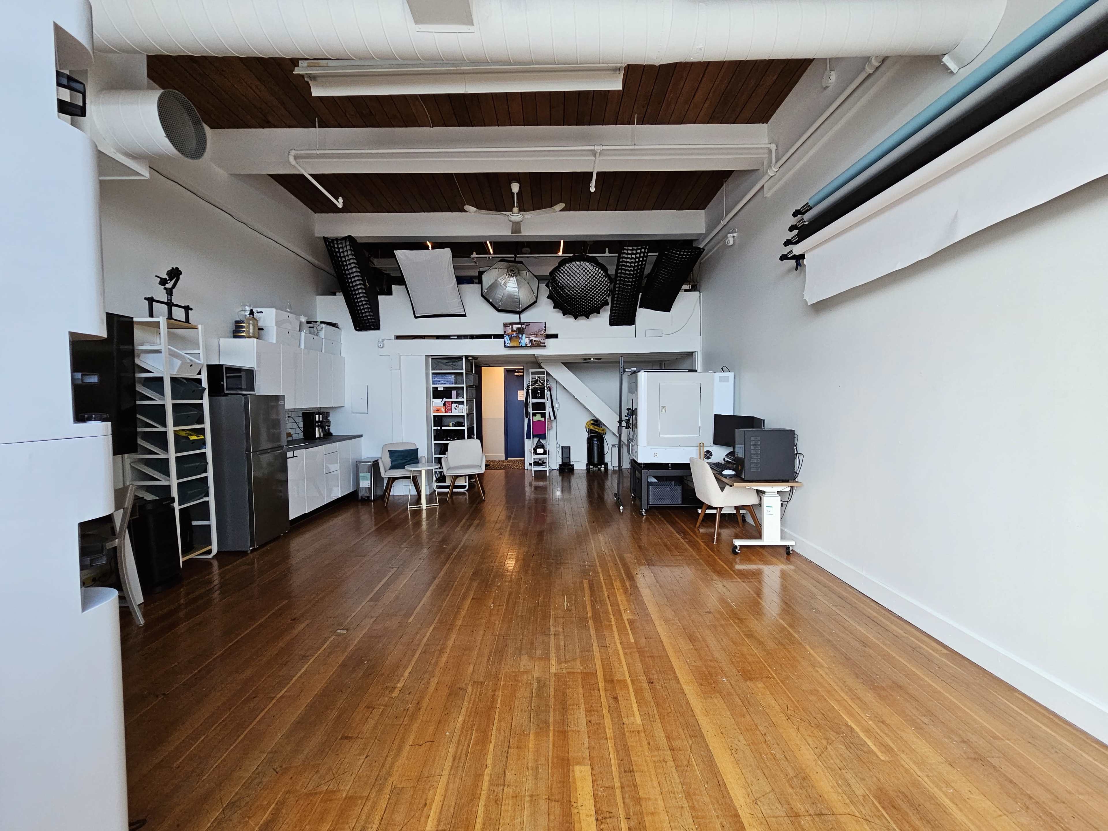 A spacious room with wooden flooring, equipped with various kitchen appliances on one side and a desk setup on the other.