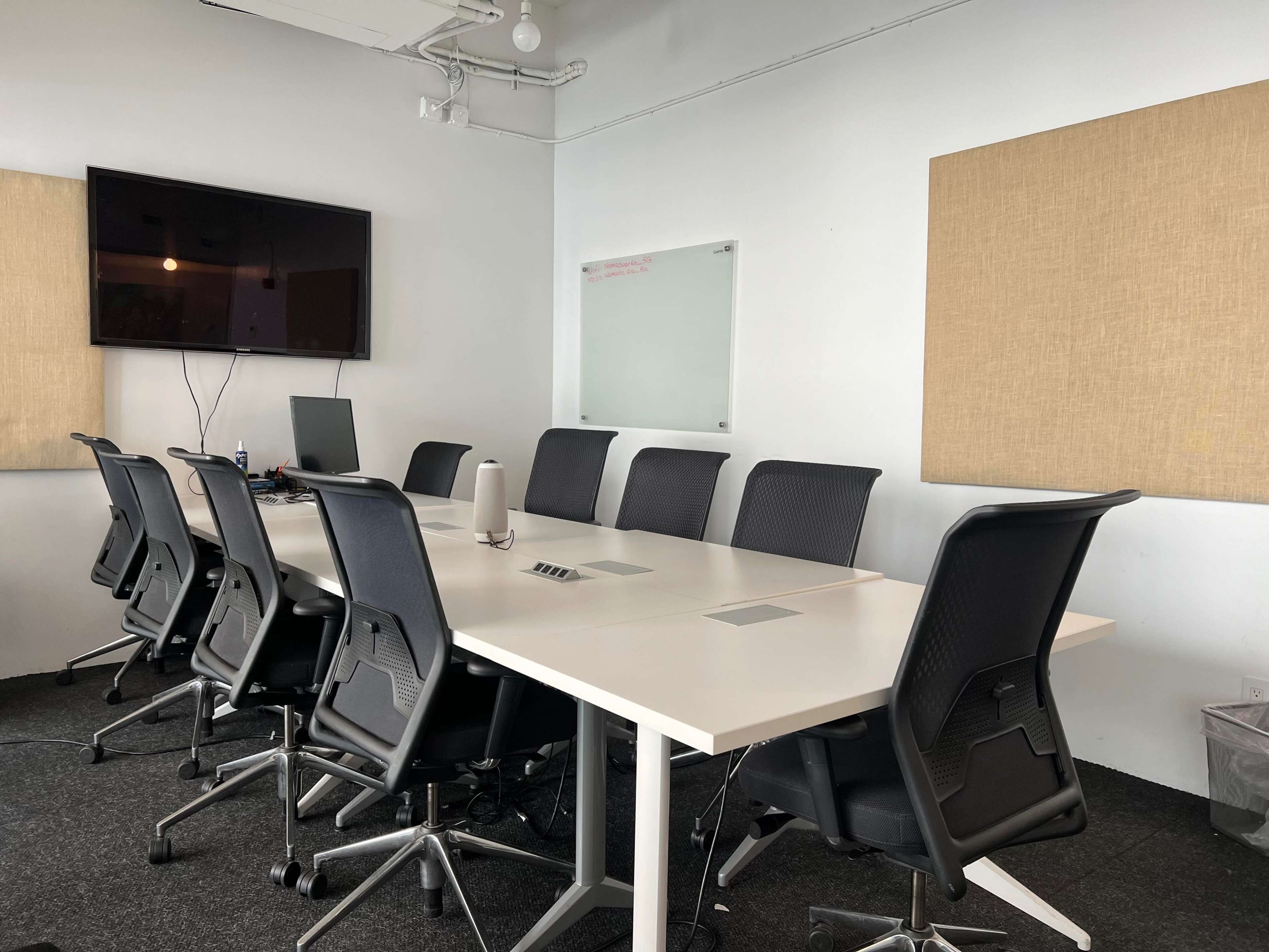 A conference room is set up with a long table, several black ergonomic chairs, a large screen on the wall, and a whiteboard.
