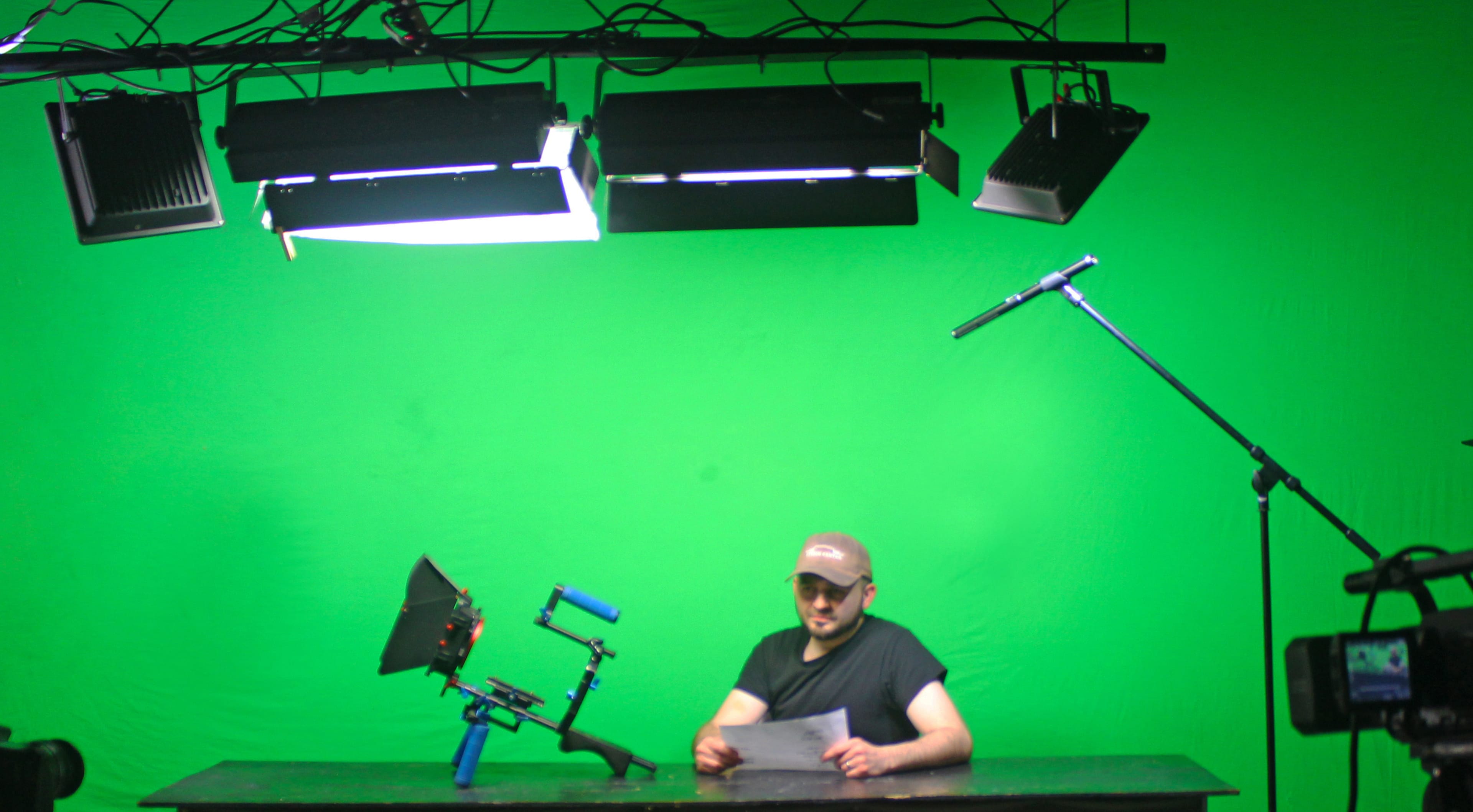 A person sits at a table in front of a green screen, holding a paper, with studio lights and a microphone positioned above them.