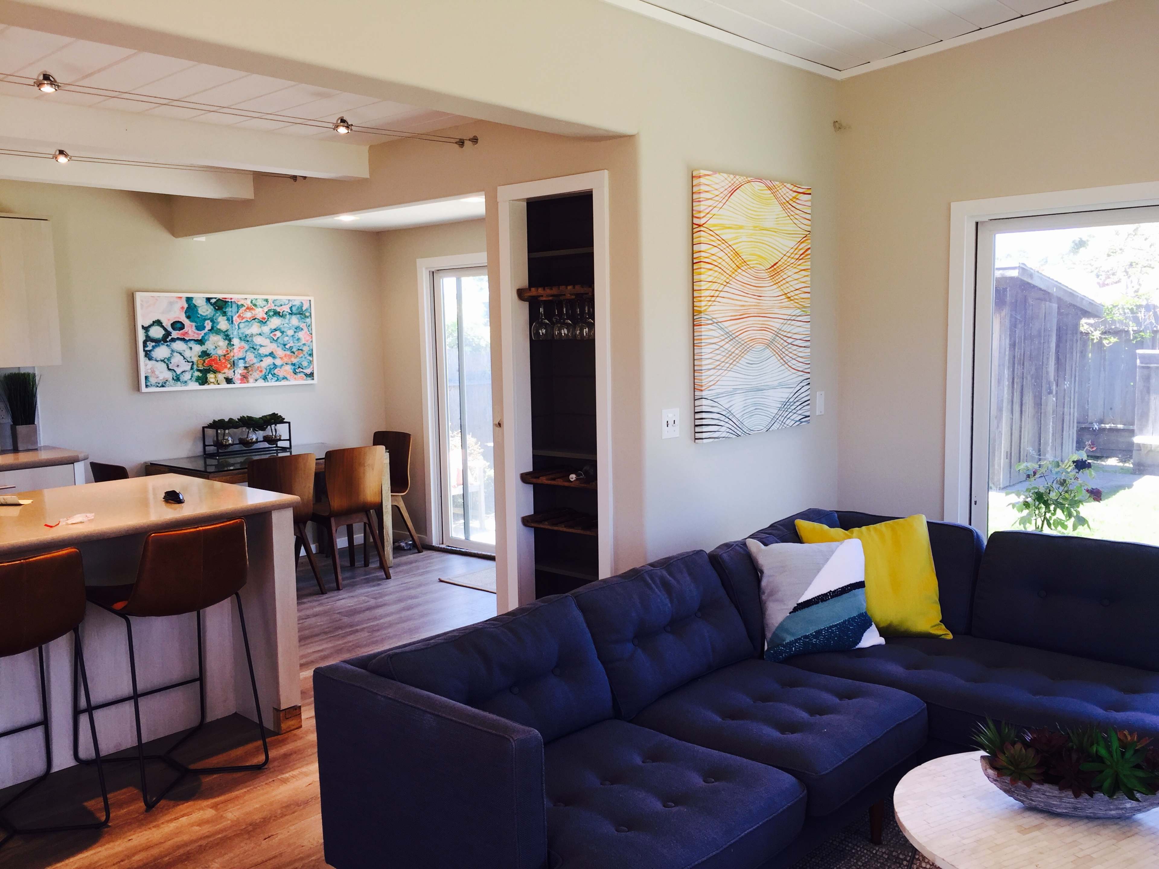 The image shows a modern living room with a sectional sofa, a round coffee table, and a dining area visible in the background, featuring large windows that allow natural light to enter.