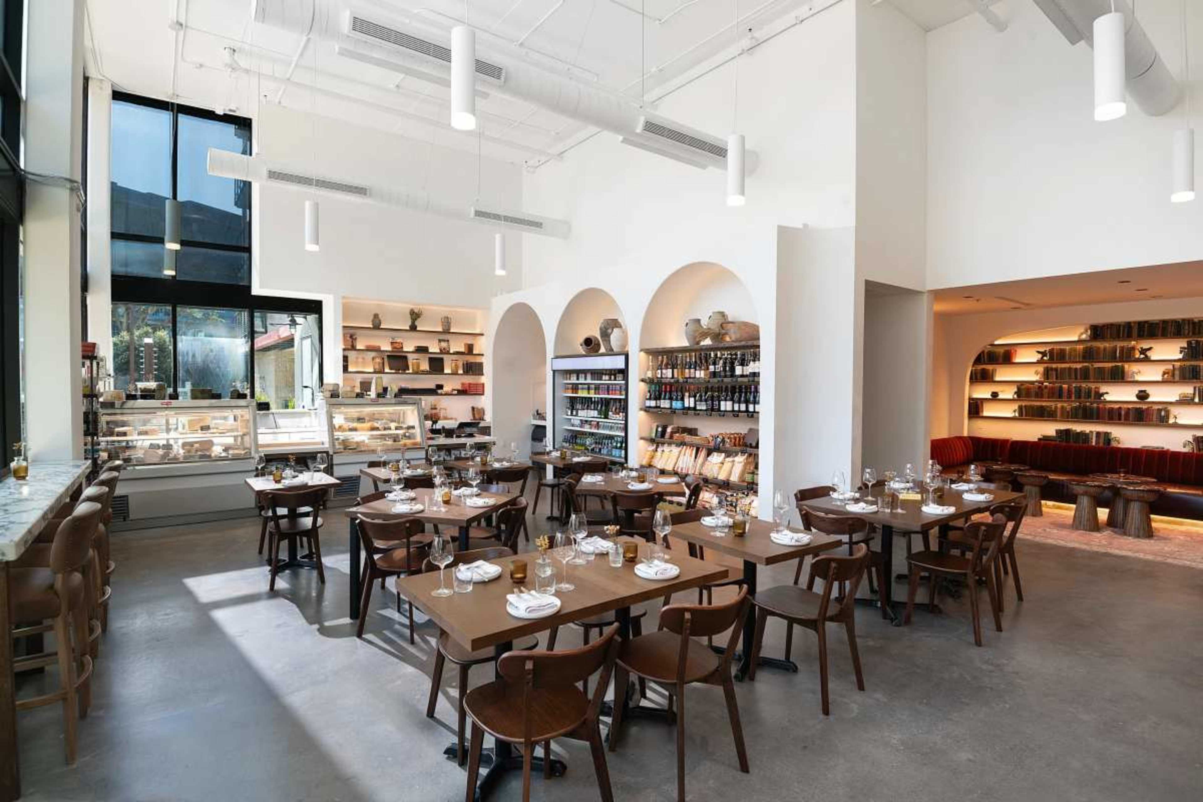 The image shows a modern restaurant interior featuring wooden tables, chairs, and shelves filled with various items, along with large windows allowing natural light to enter.