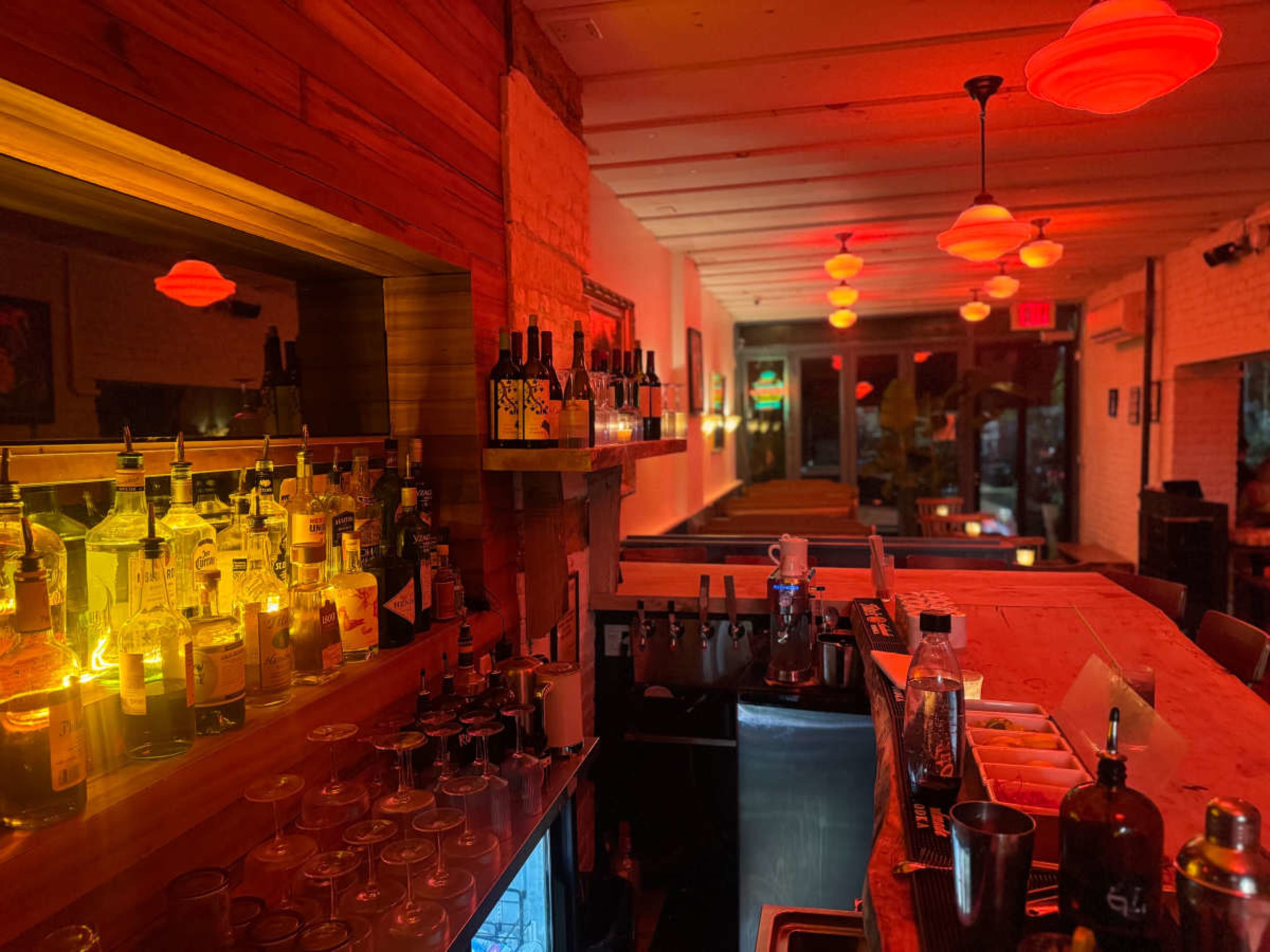 The image depicts a warmly lit bar interior featuring a variety of liquor bottles on shelves, glasses on the counter, and red pendant lights hanging from the ceiling.