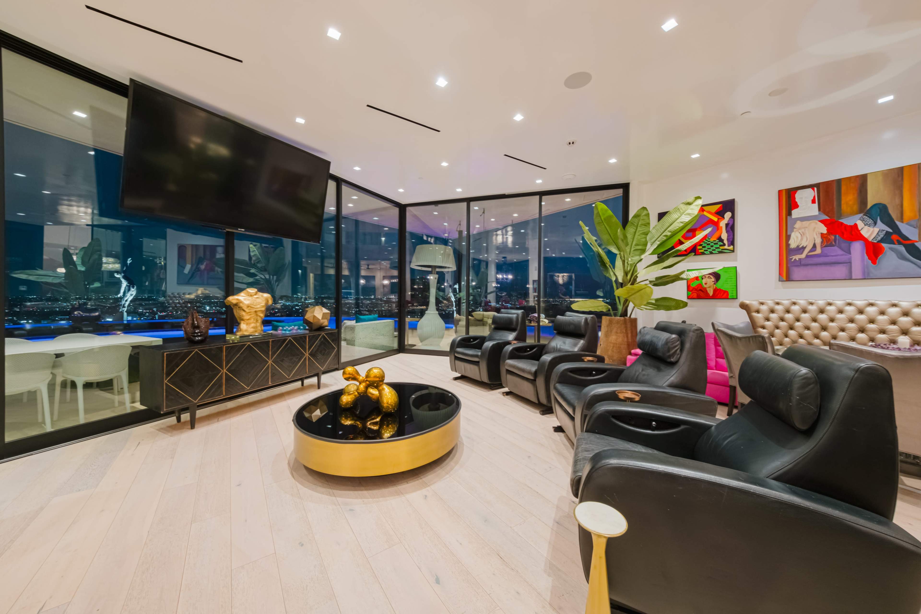 A modern living room features black leather recliners, a round coffee table with decorative gold accents, and large windows offering a view of the city at night.