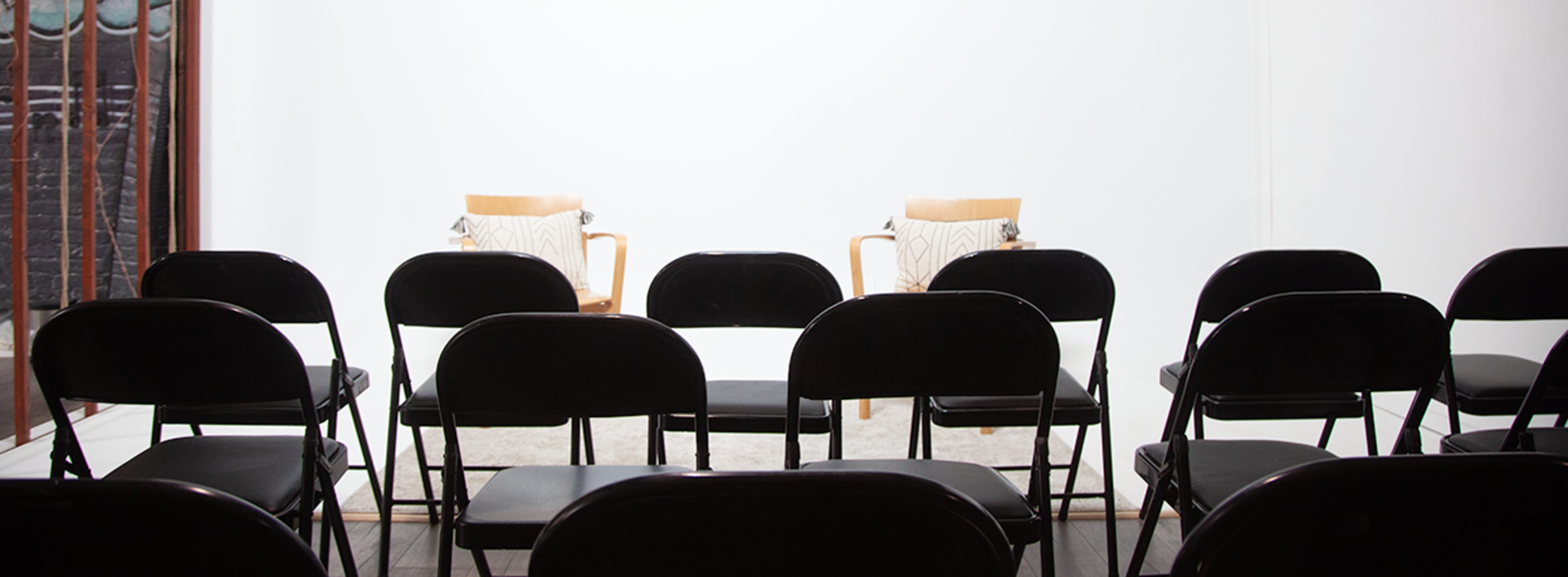 A row of black folding chairs faces a pair of wooden chairs positioned on a light-colored rug against a white wall.