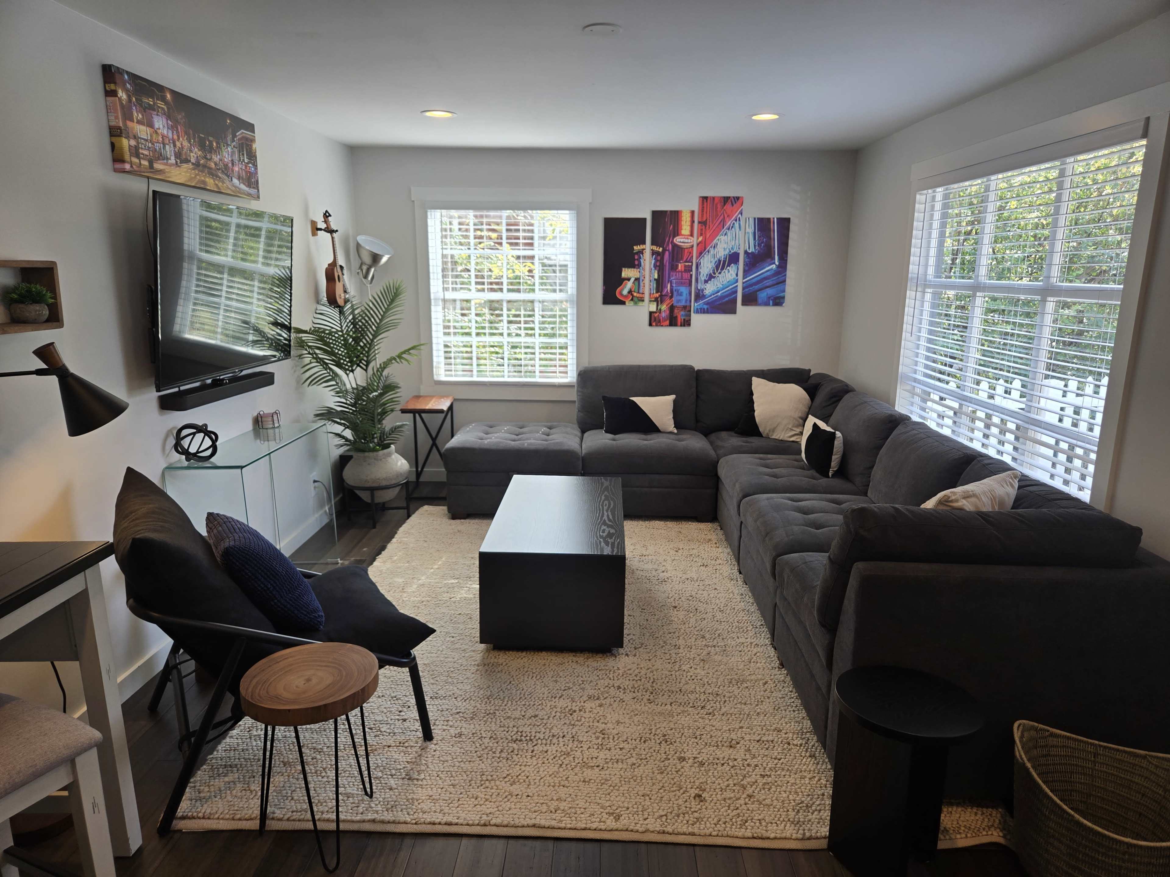 The image shows a modern living room with a dark sectional sofa, a coffee table, and colorful wall art, illuminated by natural light through multiple windows.