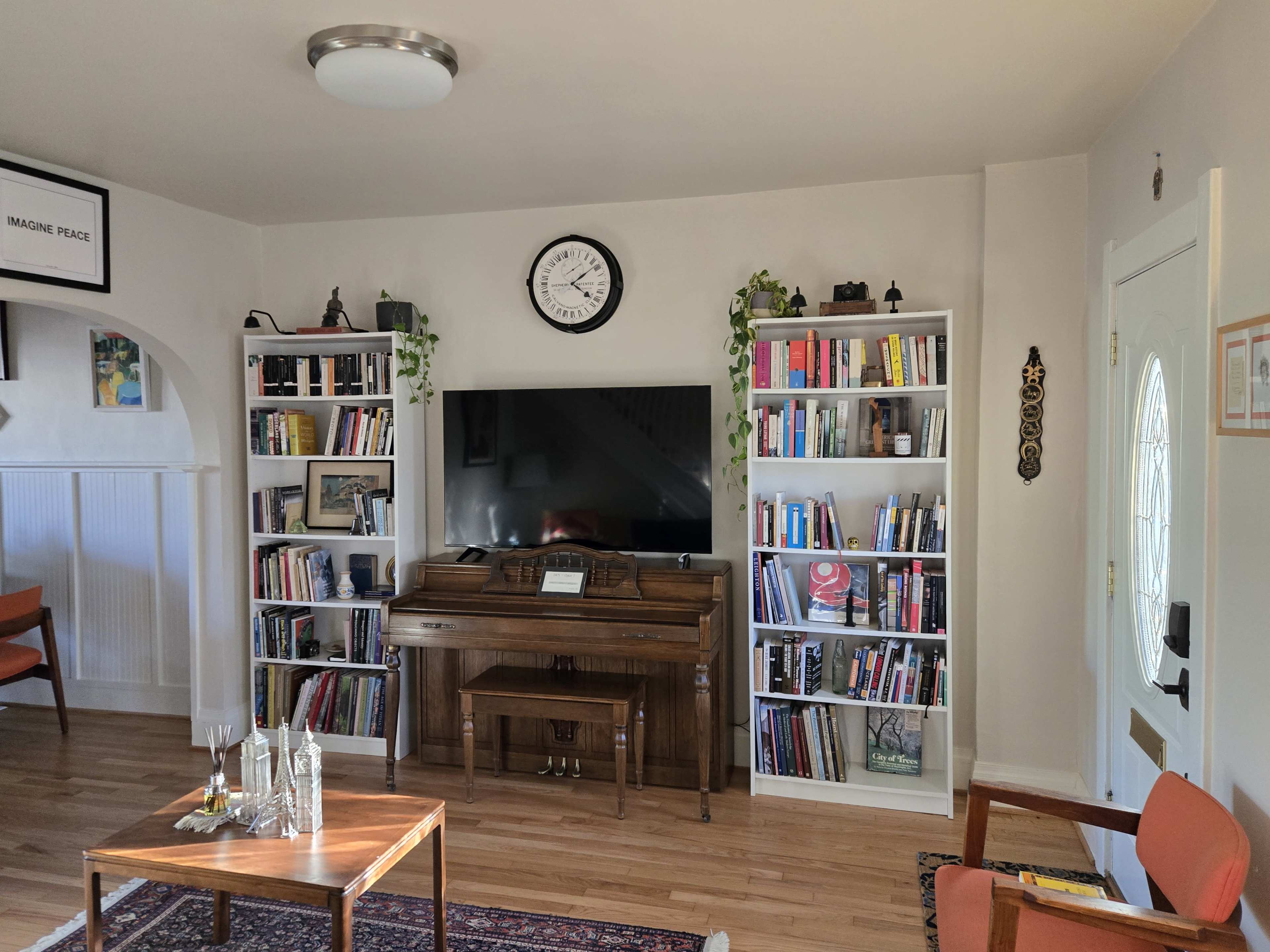 The living room features a piano between two tall bookshelves filled with books, a large television mounted on the wall, and a front door with a glass panel.