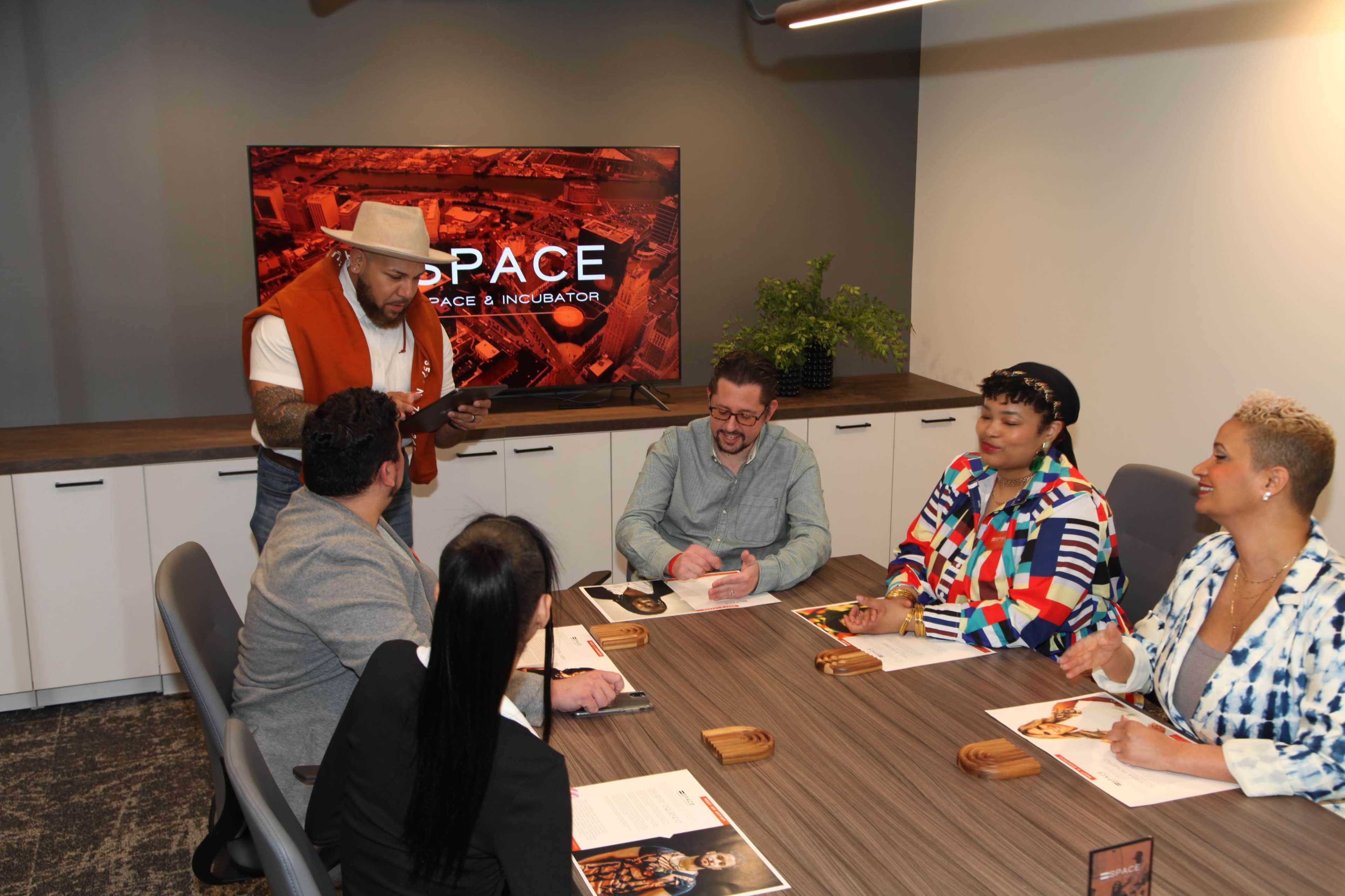 A group of five people engages in a meeting around a conference table, with a large screen displaying "SPACE" in the background.