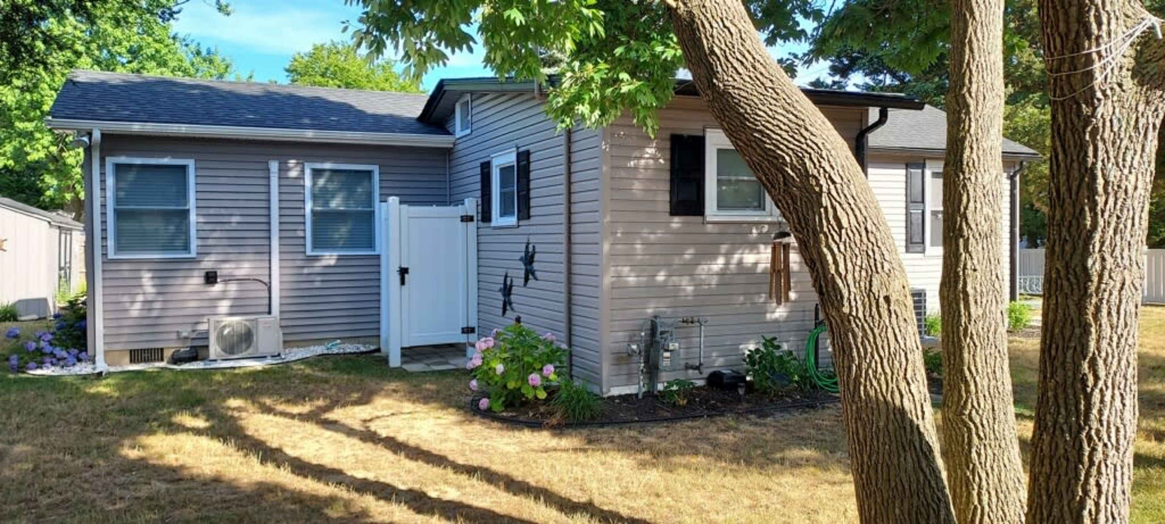 A single-story house with light-colored siding, a white fence, and landscaping featuring grass and bushes in a residential area.