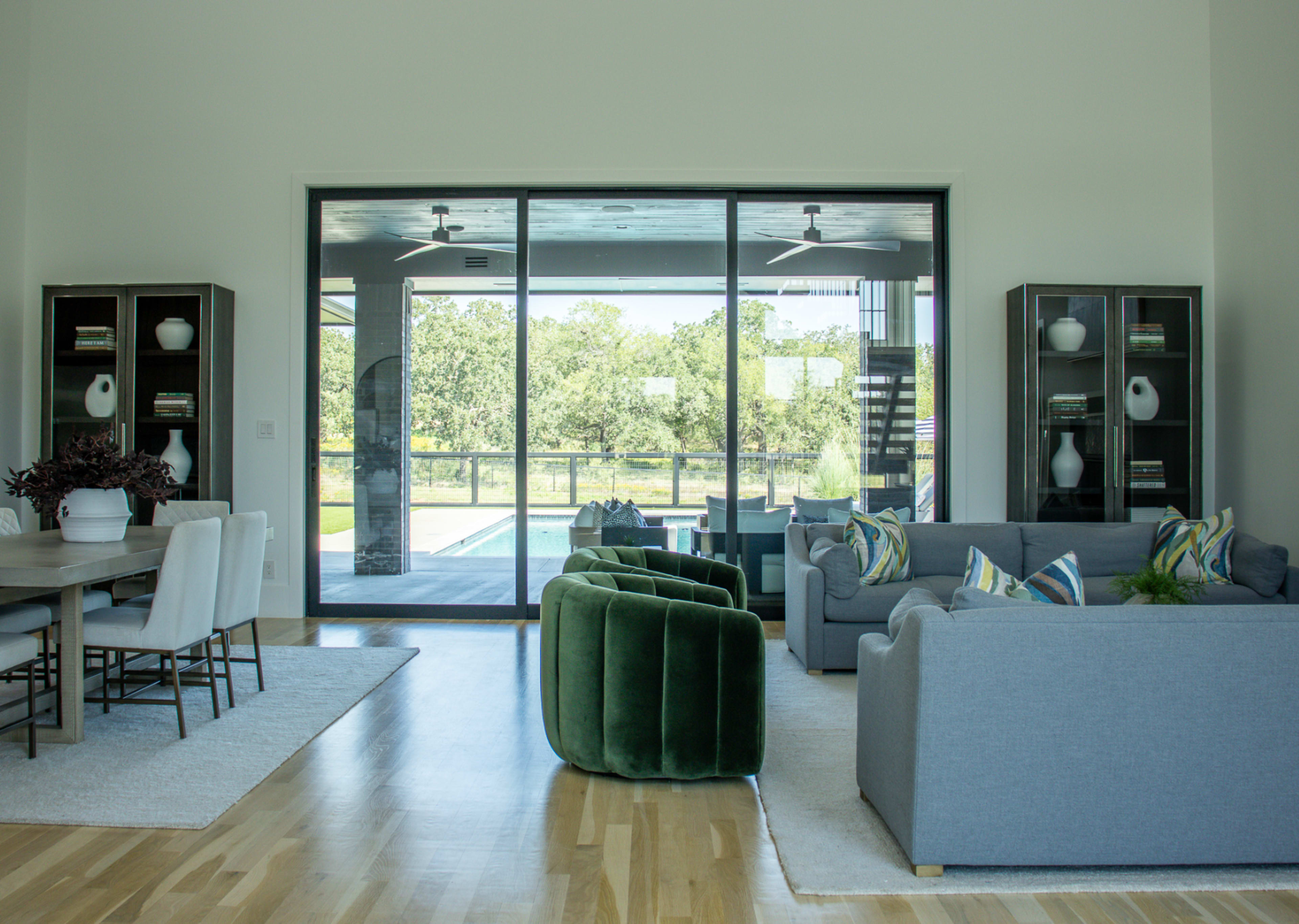 A modern living room features a green armchair and gray sofas, with a dining table nearby and large windows overlooking a pool and outdoor landscape.