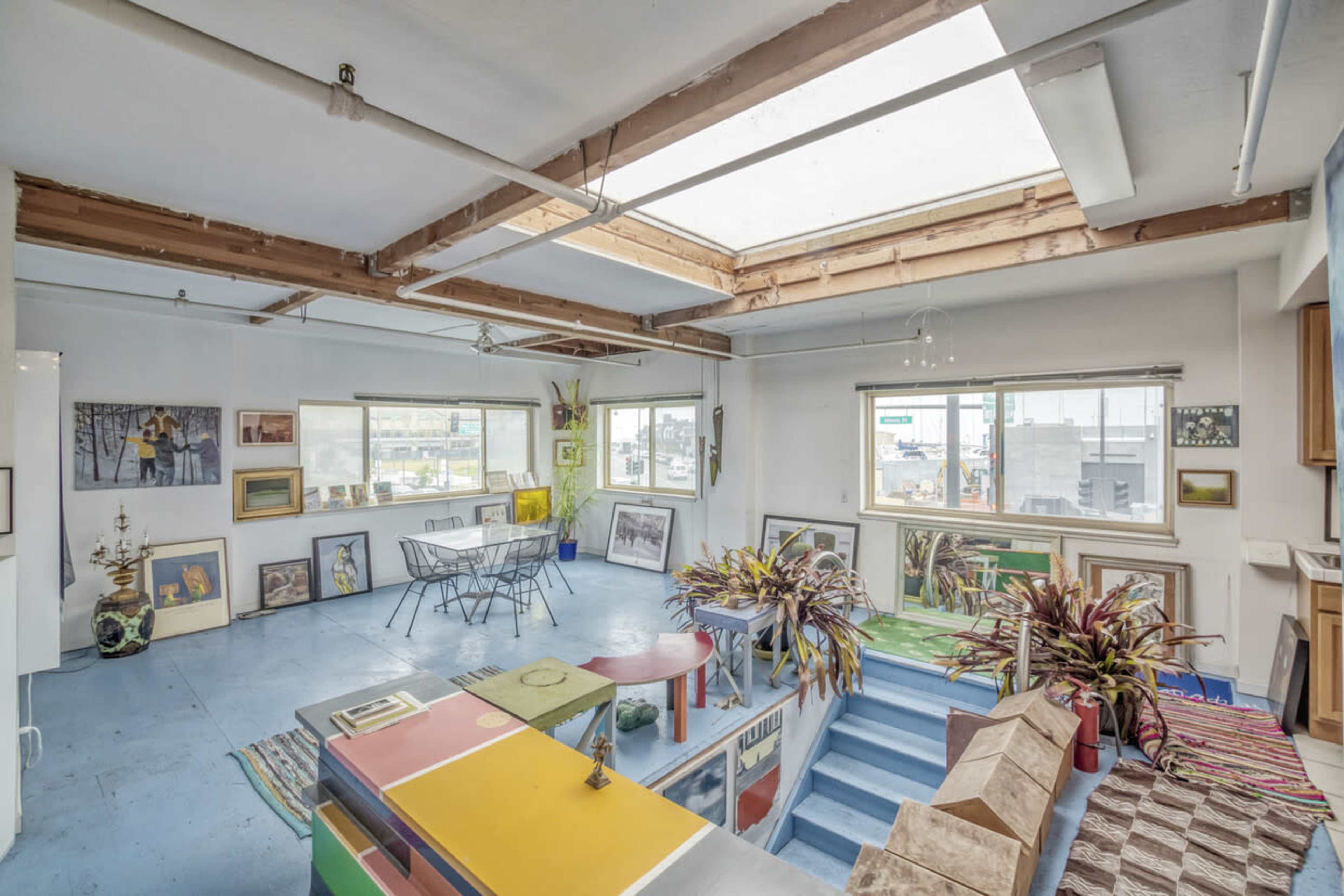 A spacious, light-filled room featuring an open layout with colorful furniture, numerous framed artworks on the walls, and large windows.