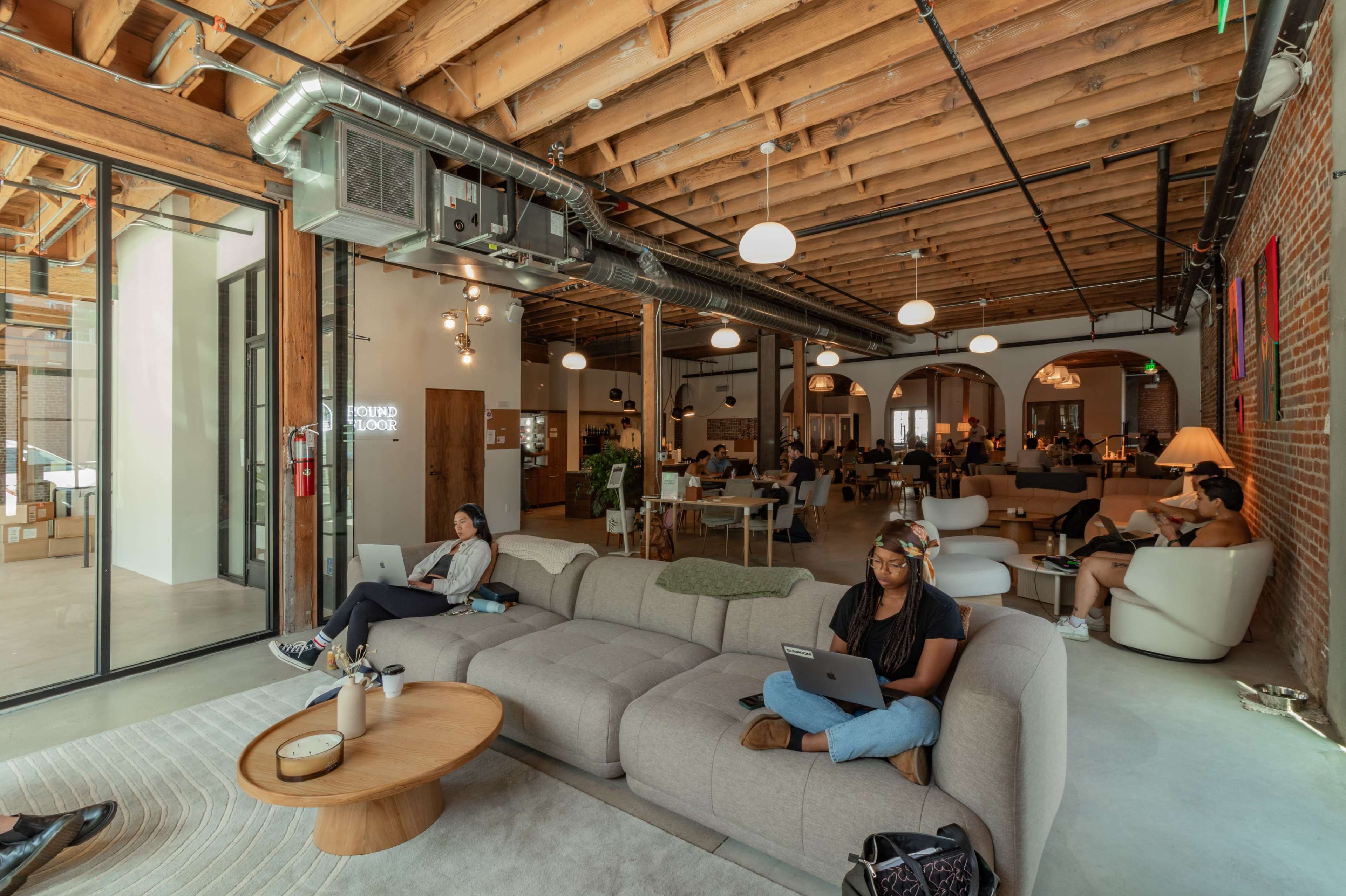 A modern workspace features a large, light-colored sectional sofa with people using laptops, surrounded by an open area filled with varied seating and wooden ceiling beams.