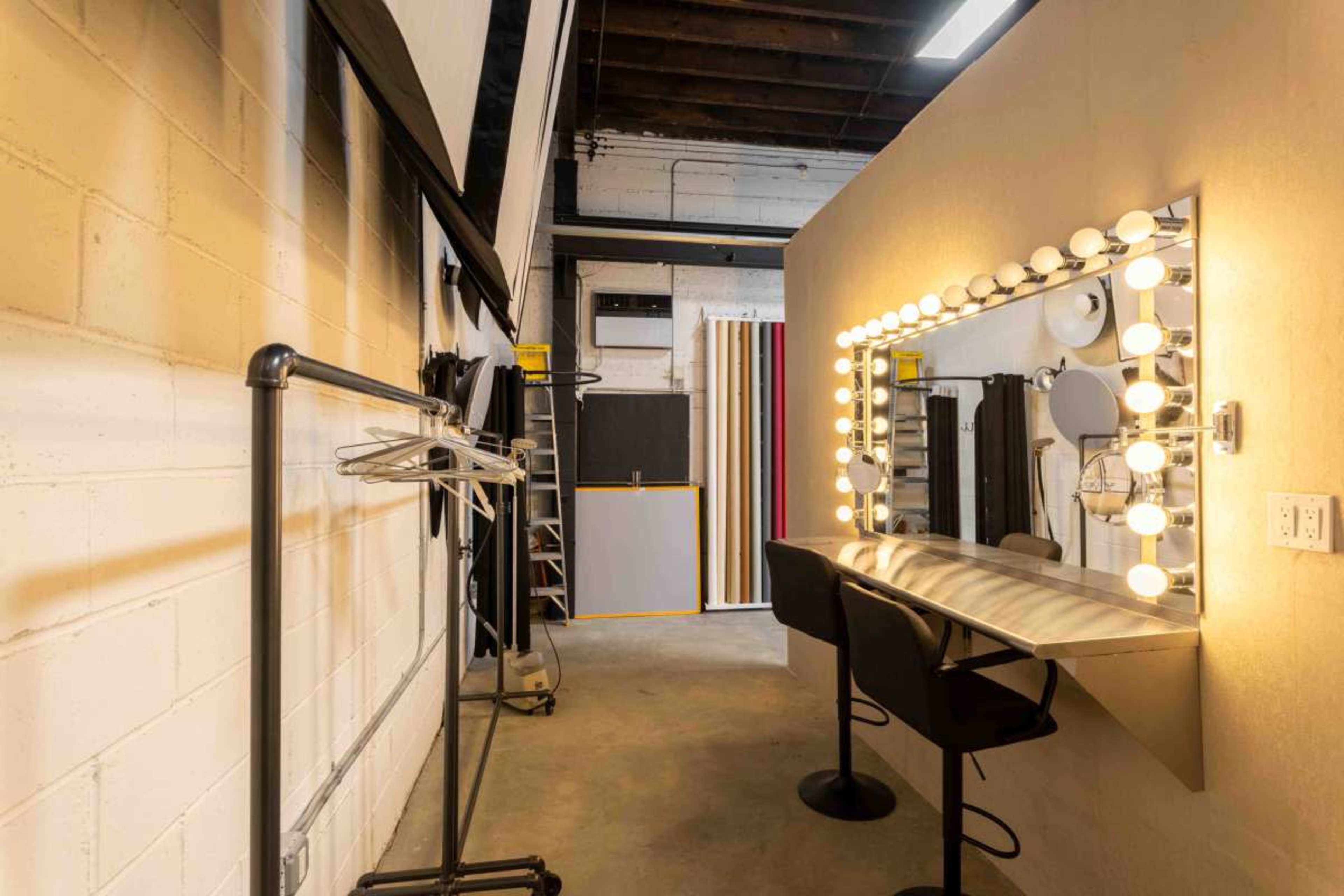 The image shows a makeup room with a large mirror surrounded by light bulbs, two black stools, and empty clothing racks against white brick walls.