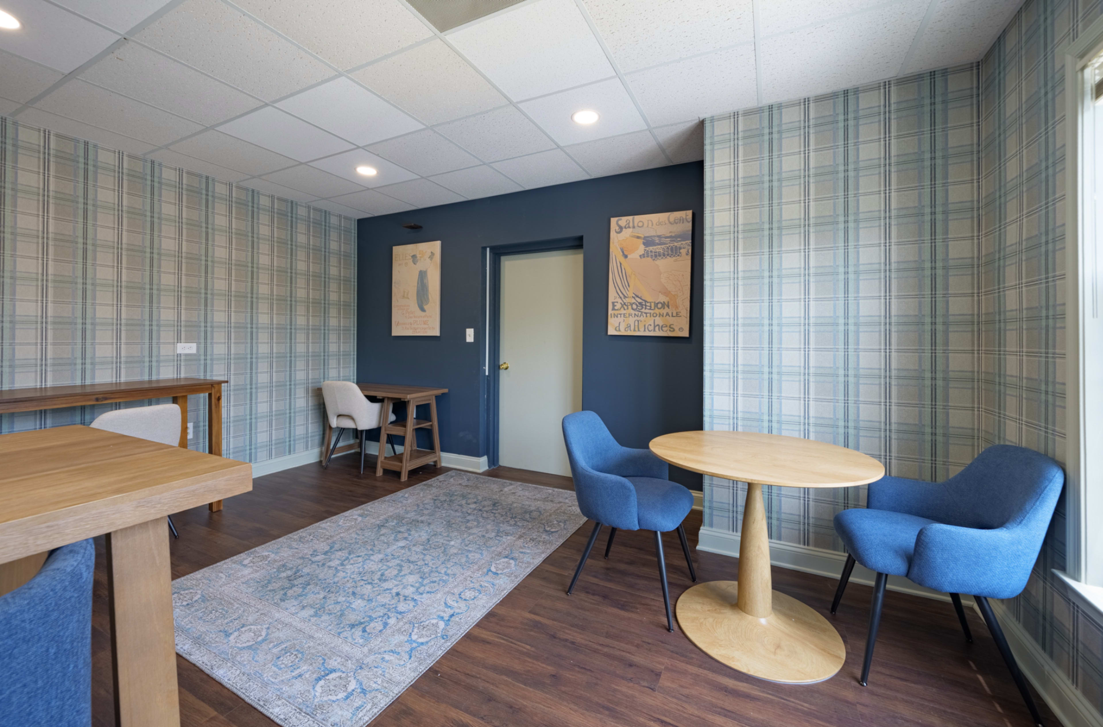 The image depicts a modern, indoor space featuring a small round table with two blue chairs, a wooden table, and a patterned wallpapered wall.