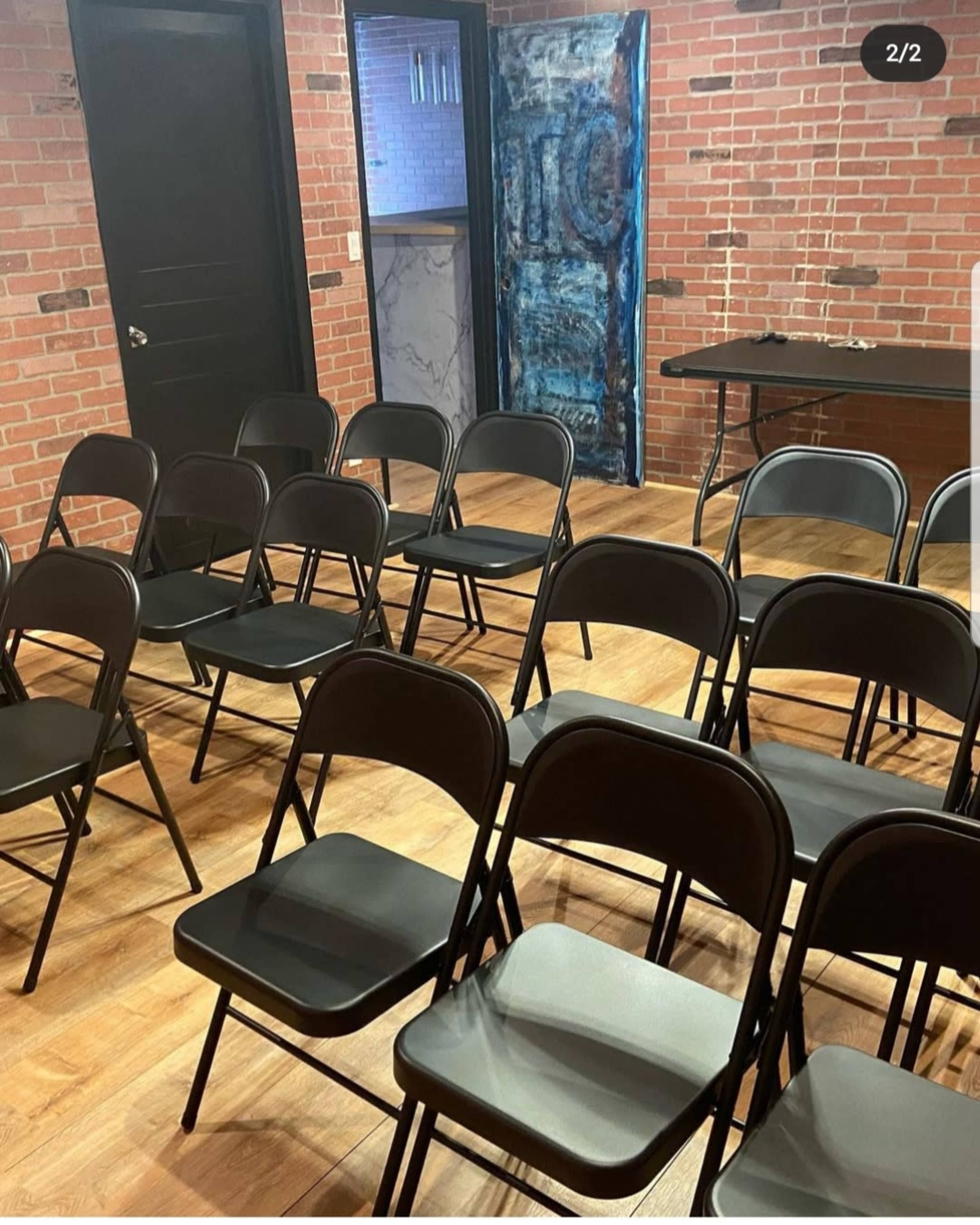 A room with several black folding chairs arranged in rows on a wooden floor, with brick walls and a table in the background.