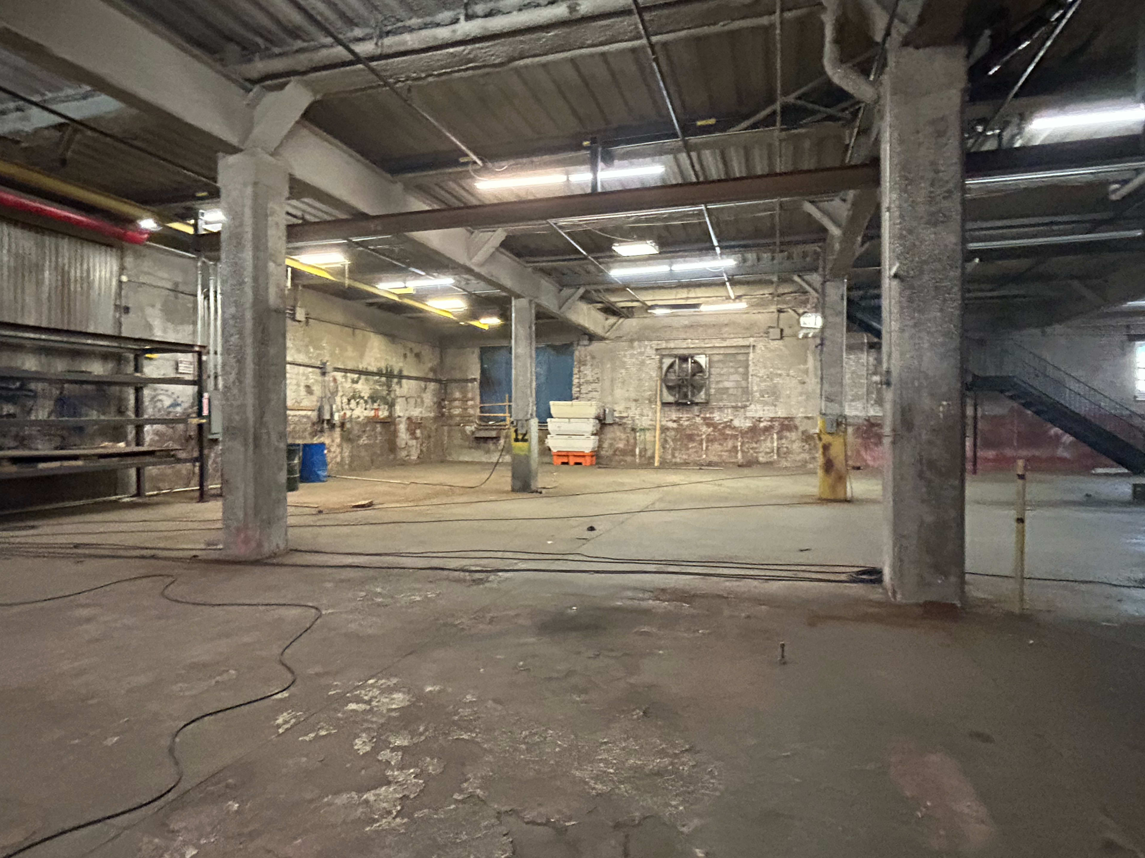 The image shows a spacious, industrial interior with concrete walls, exposed beams, and minimal furnishings, including barrels and a staircase in the background.