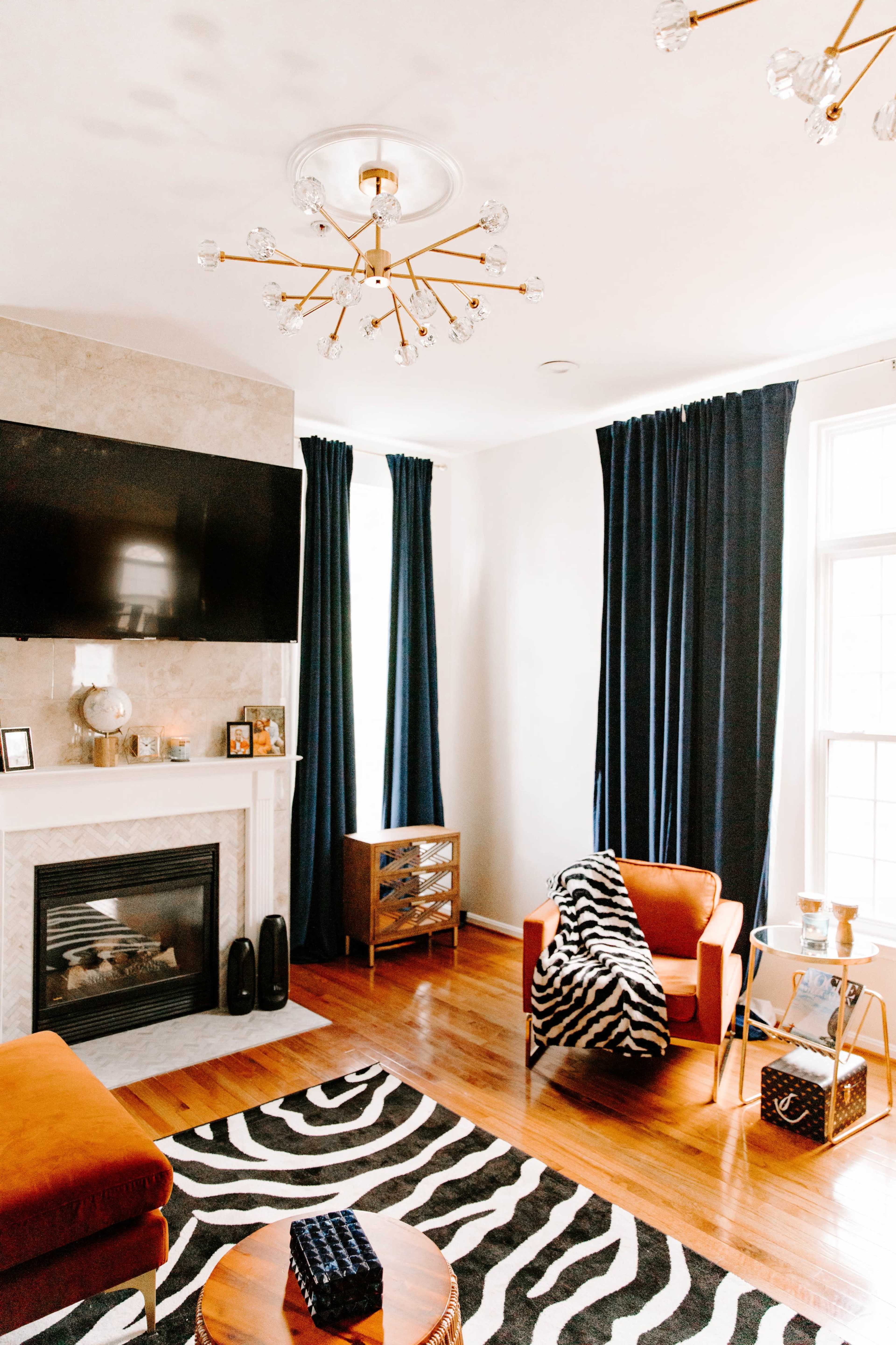The living room features a fireplace, a television mounted above the mantle, orange armchairs, a zebra-patterned rug, and navy curtains.