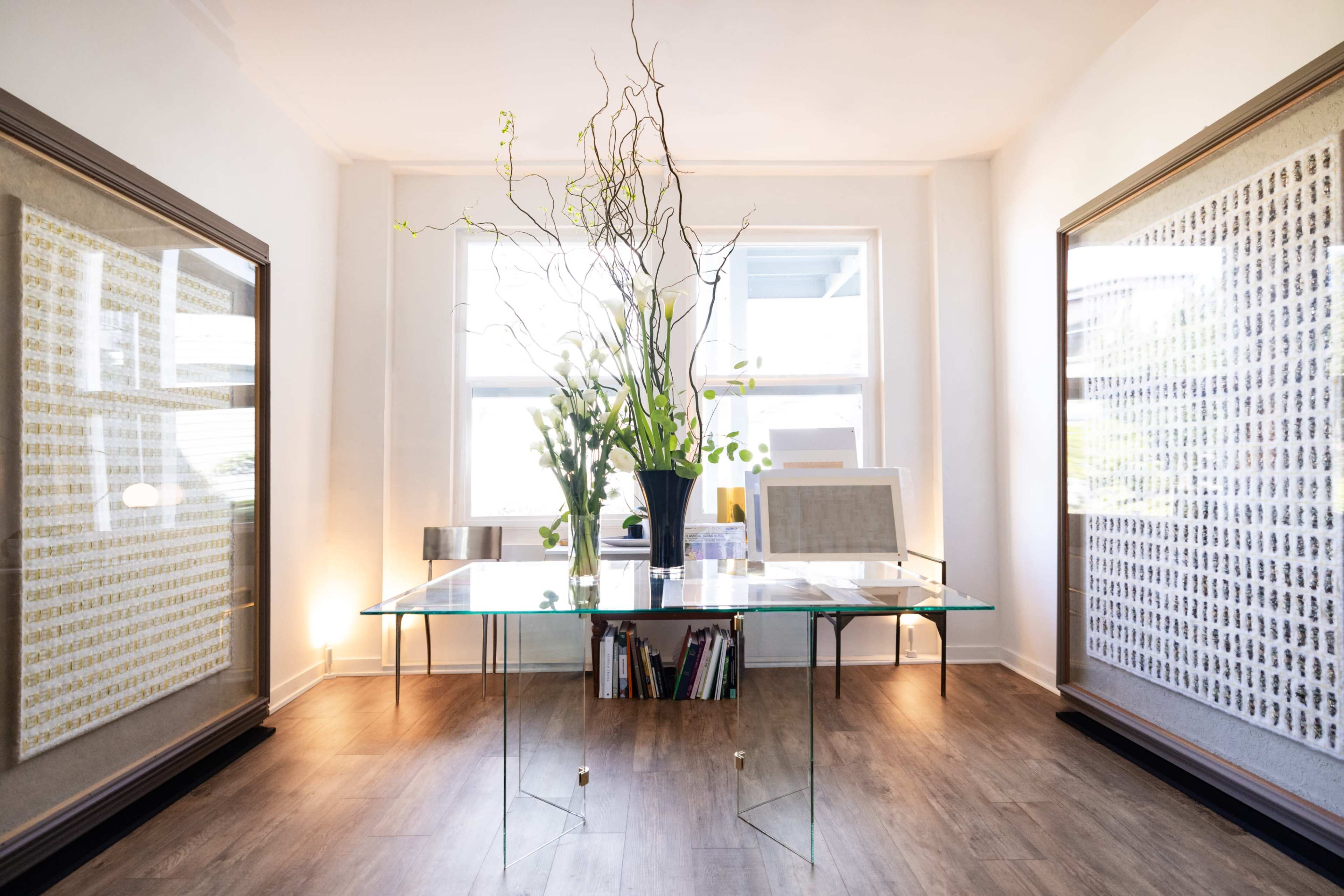 A modern office space with a glass table, surrounded by large framed artworks, and a large floral arrangement in a vase.