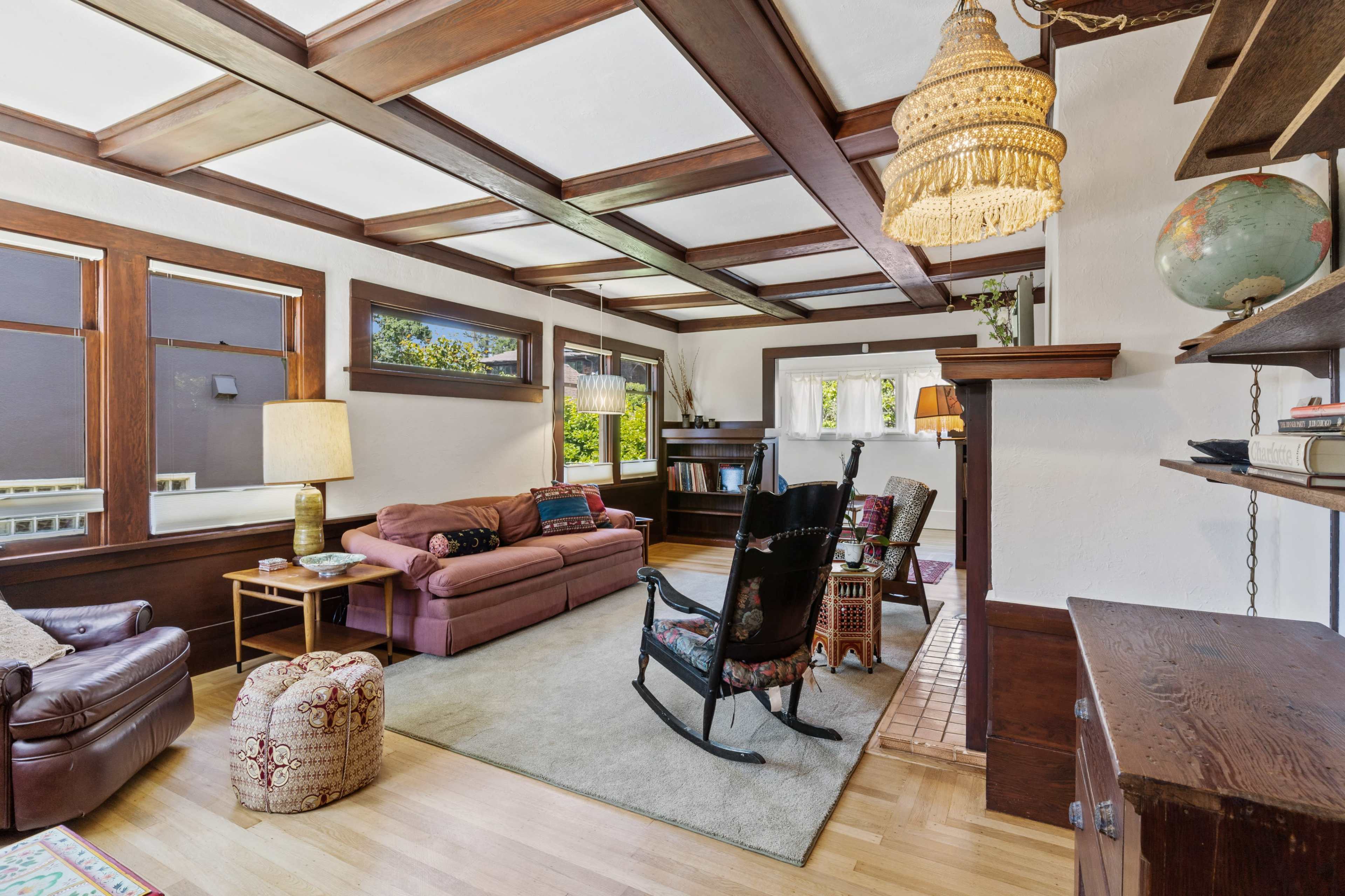 A living room features a large sectional sofa, a rocking chair, and wooden beams on the ceiling, with bookshelves and windows allowing natural light.
