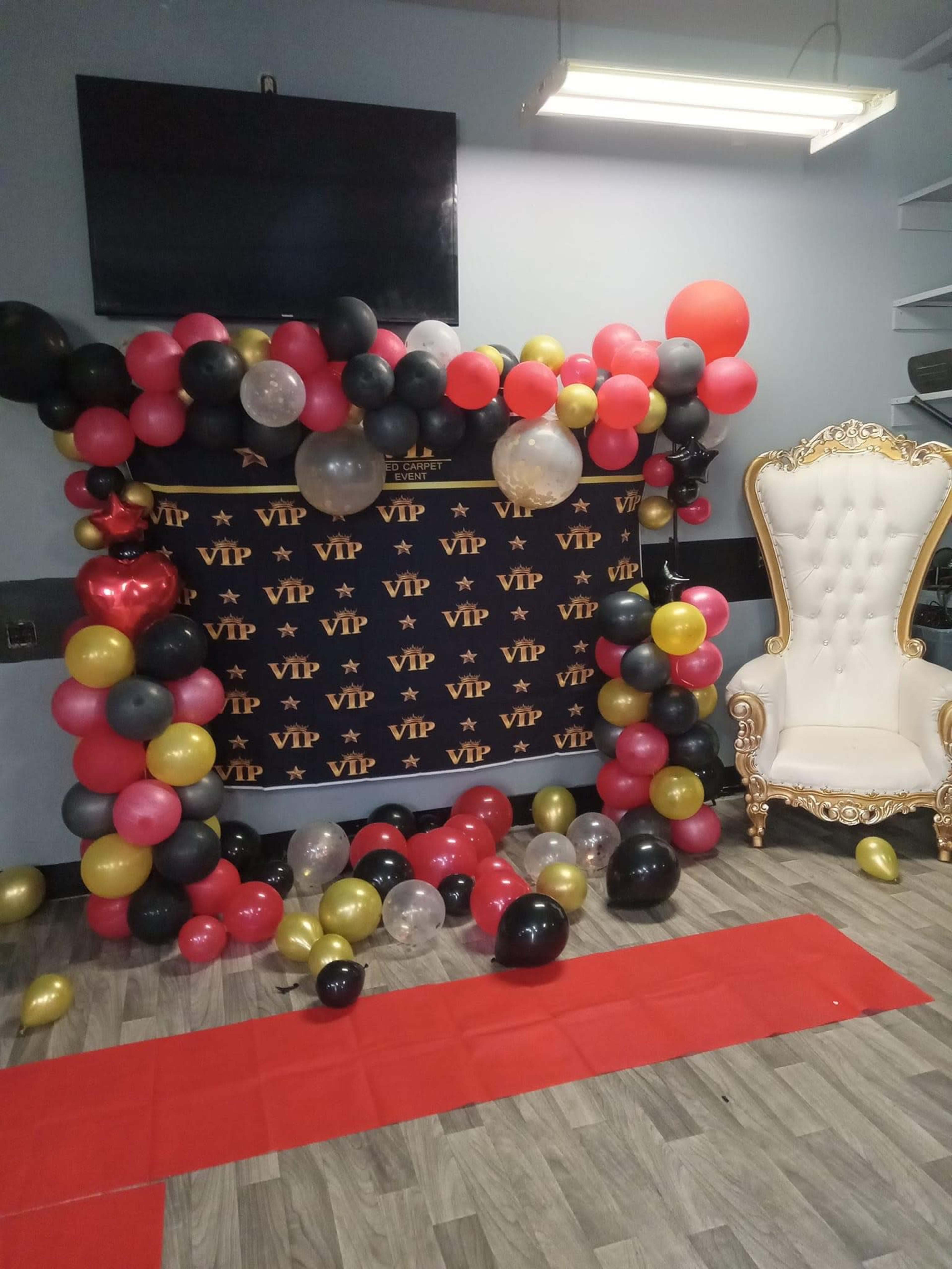 The image shows a decorated area featuring a balloon arch in black, red, gold, and silver, with a white throne-like chair and a red carpet leading towards a backdrop that reads "VIP."