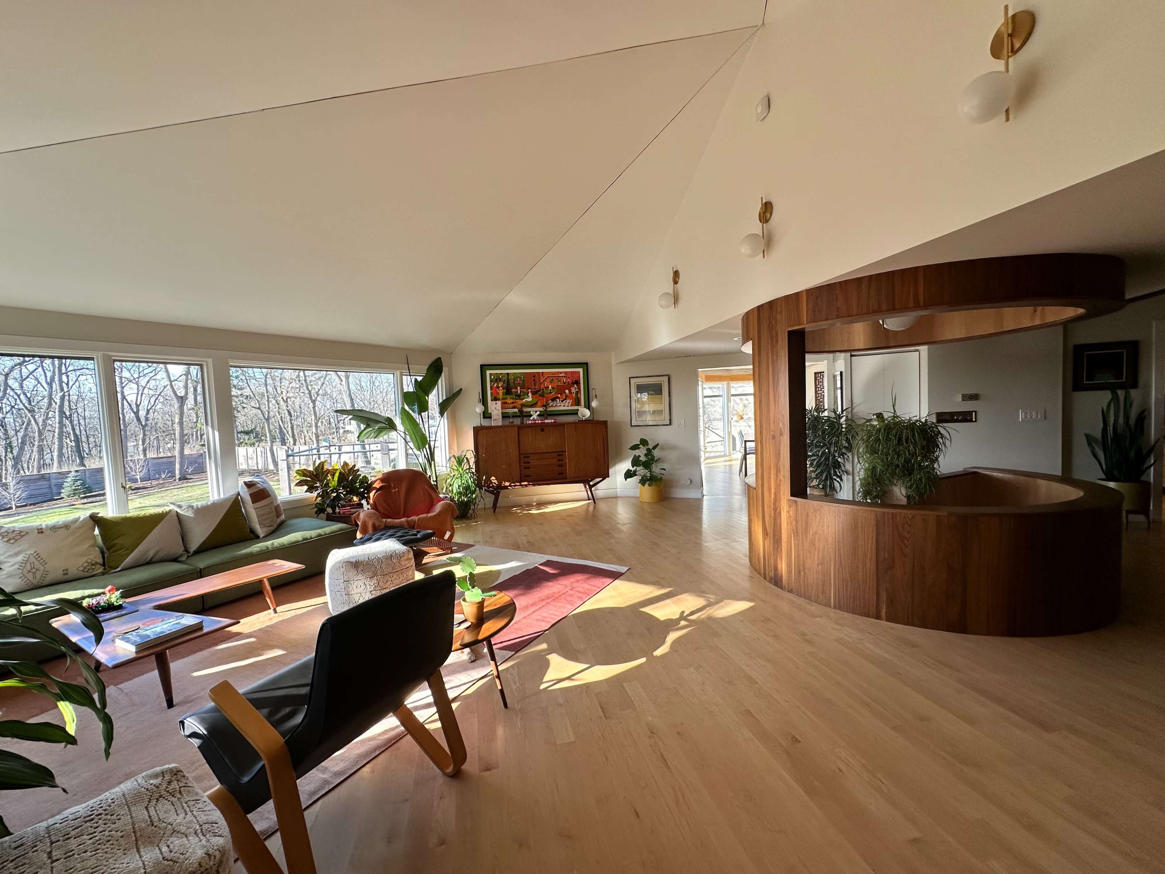 A spacious modern living room with large windows, a wooden central structure, and various plants and furniture arranged around the space.