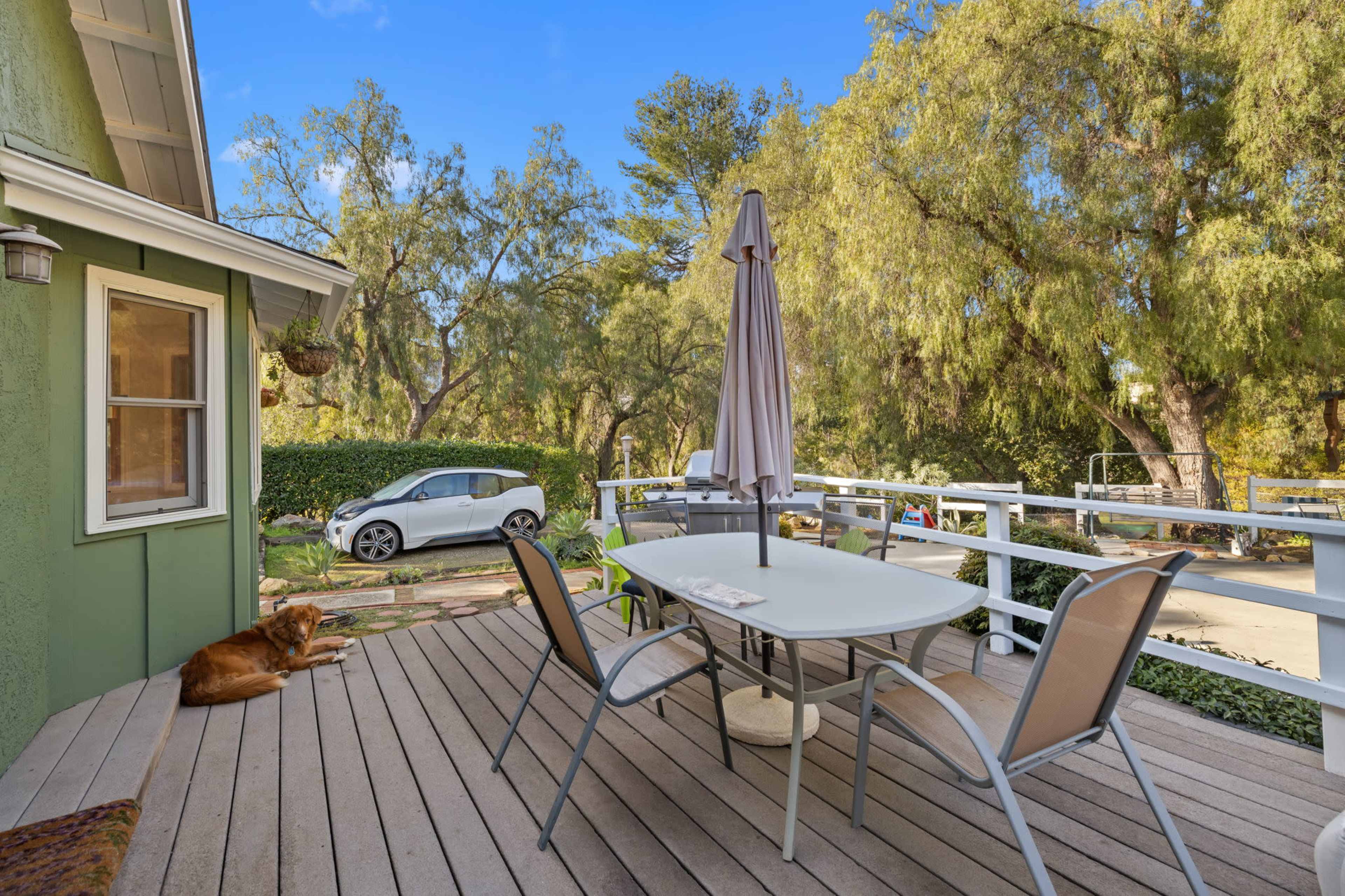 A patio with a table, chairs, and an umbrella overlooks a green landscape, while a dog rests nearby and a car is parked in the driveway.