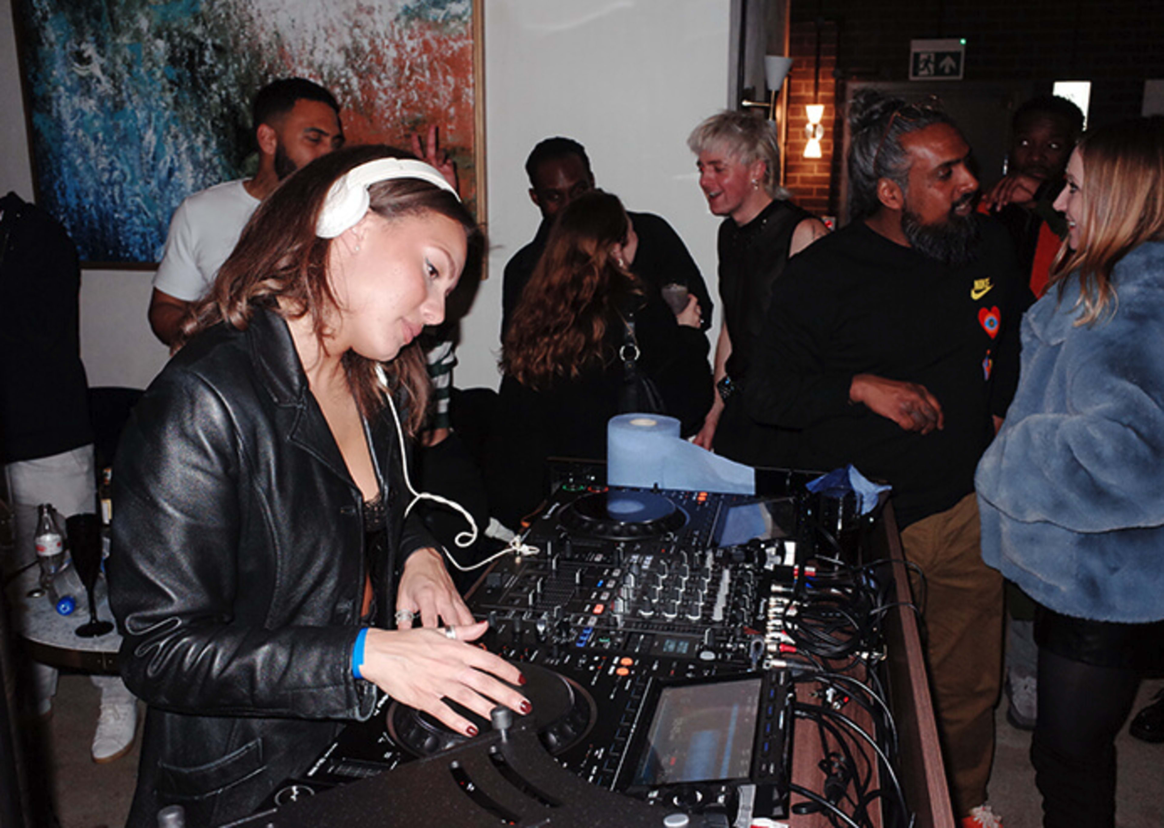 A female DJ is focused on her equipment while a diverse group of people socializes around her in a lively indoor setting.