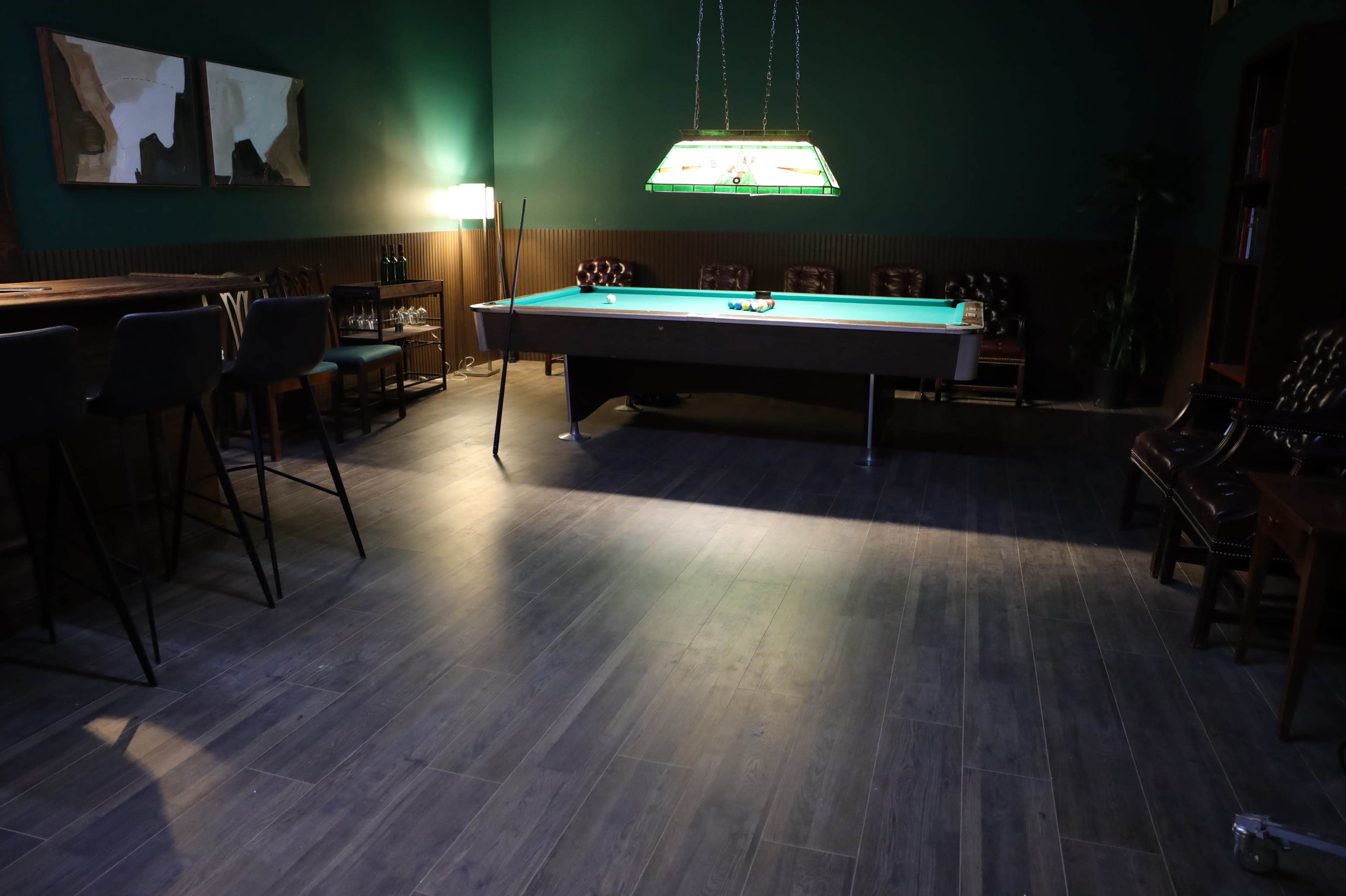 The image shows a dimly lit room with a green pool table at the center, surrounded by chairs and tables, and a bar area in the background.