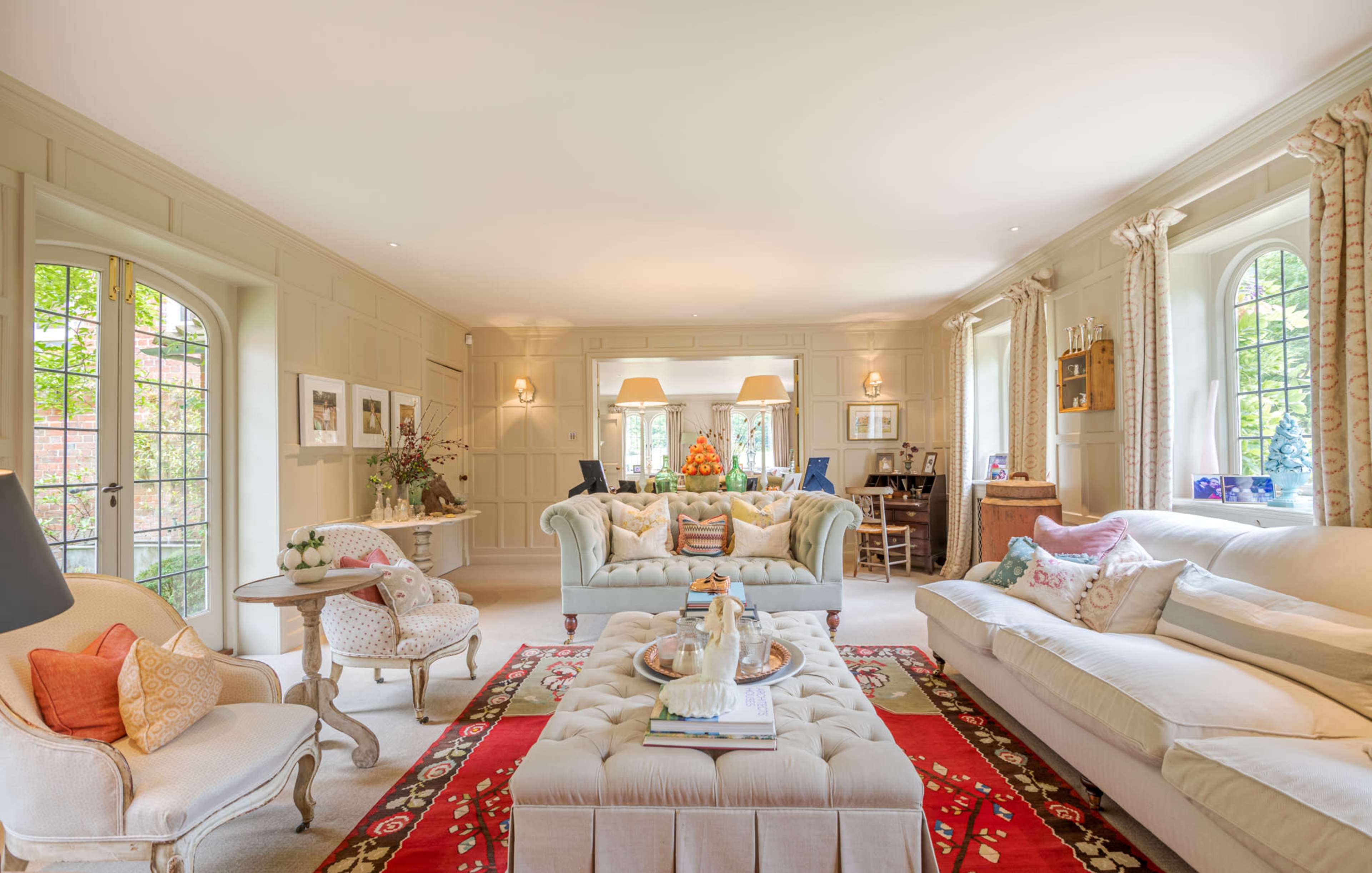 The image shows a spacious living room furnished with a light-colored sofa, a tufted ottoman, and large windows that allow natural light to fill the space.