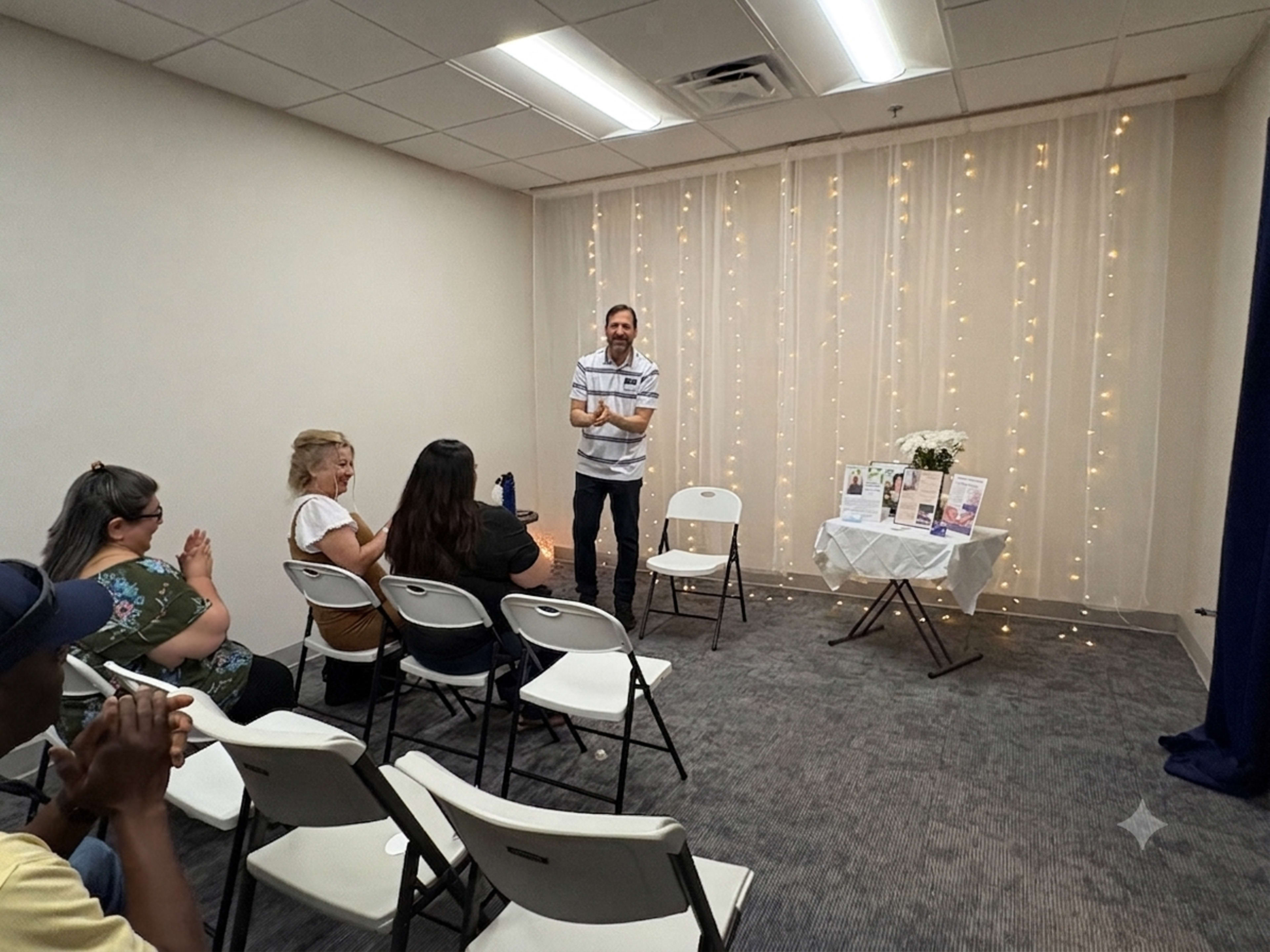 A man stands in front of a small group of seated individuals in a brightly lit room decorated with string lights and a table displaying various items.