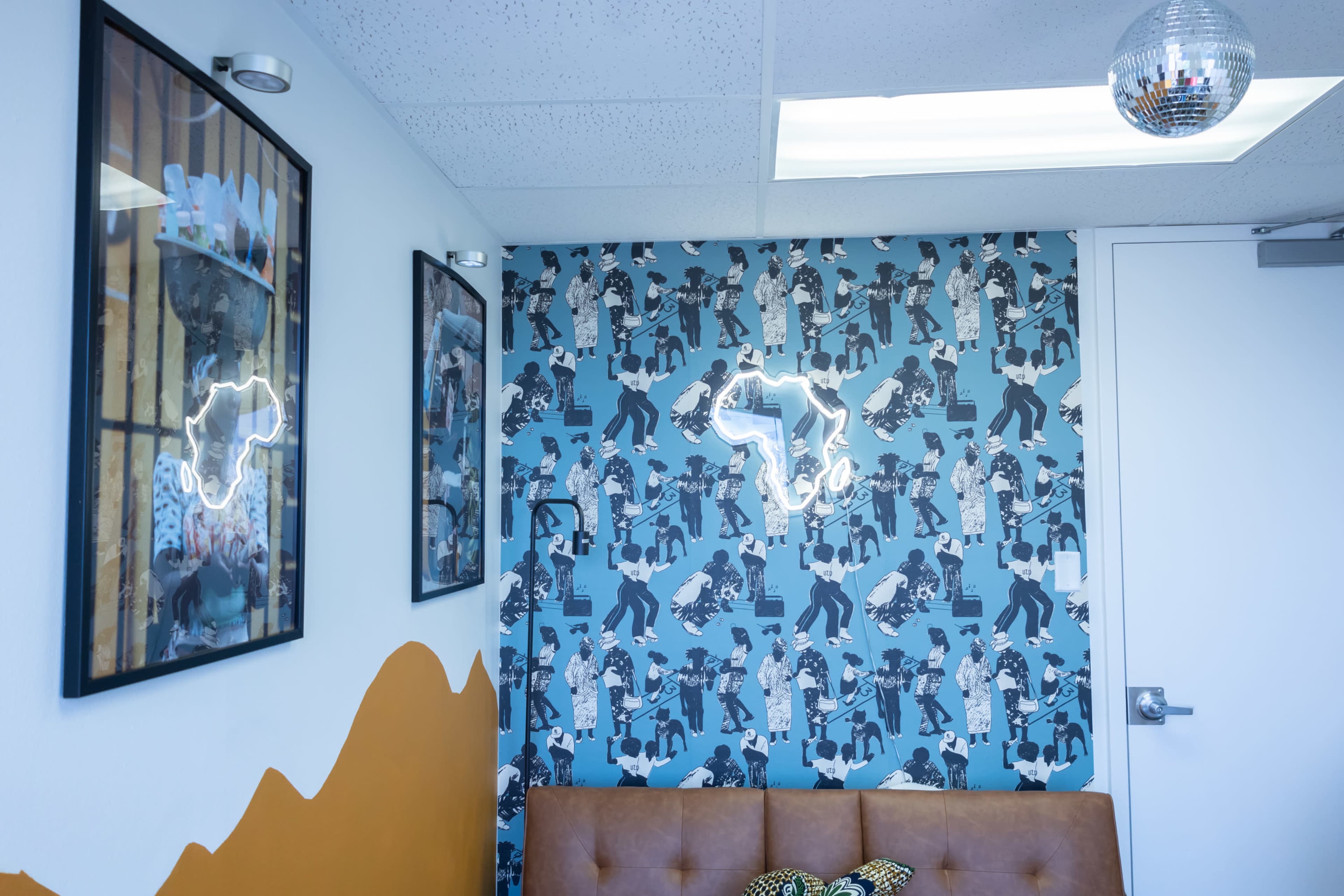 A corner of a room featuring patterned wallpaper with a light fixture in the shape of Africa, framed artwork on the walls, and a brown sofa.