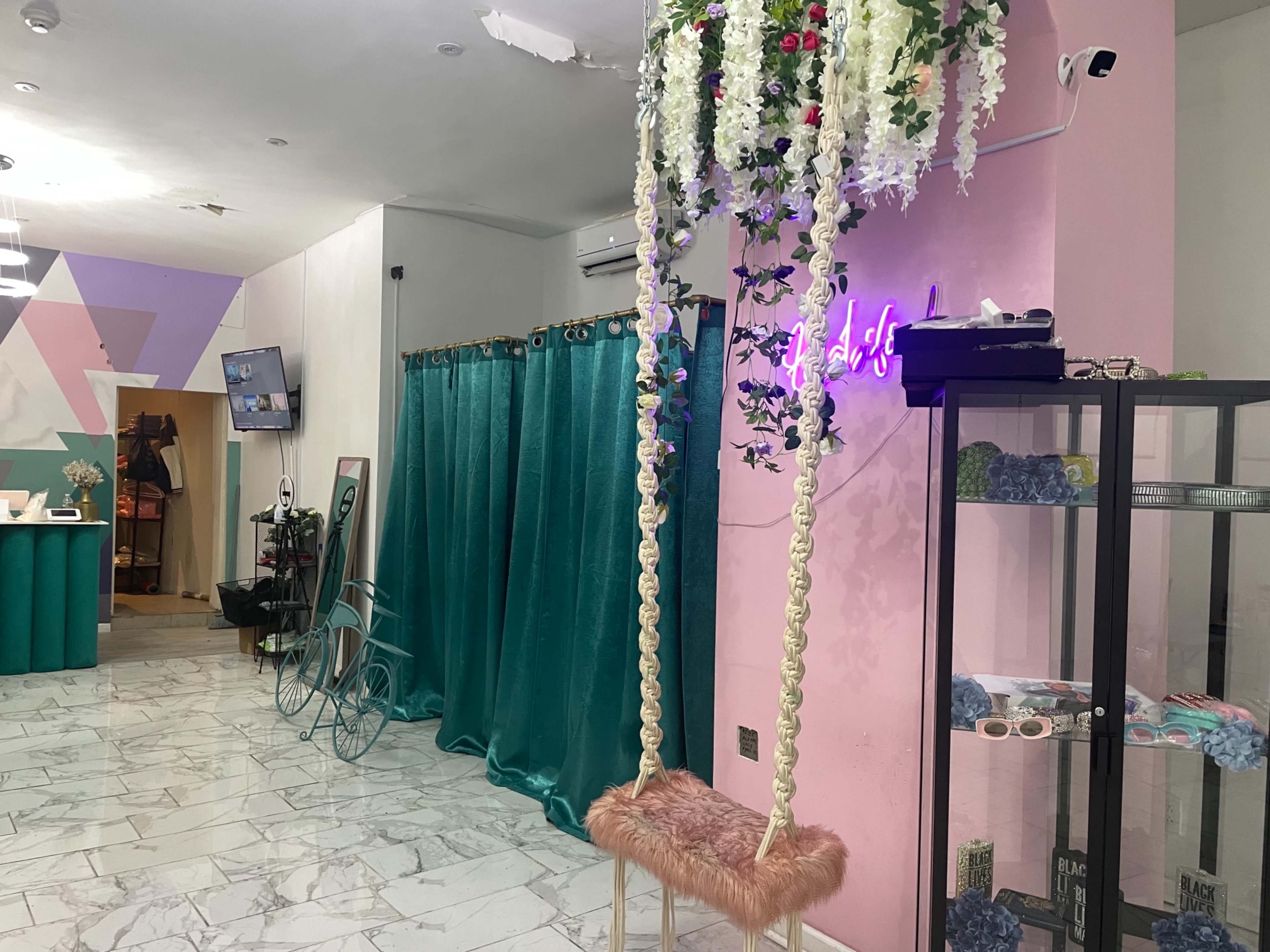 A boutique interior with a pink wall, green curtains, a swing adorned with flowers, and display cases showcasing various items.