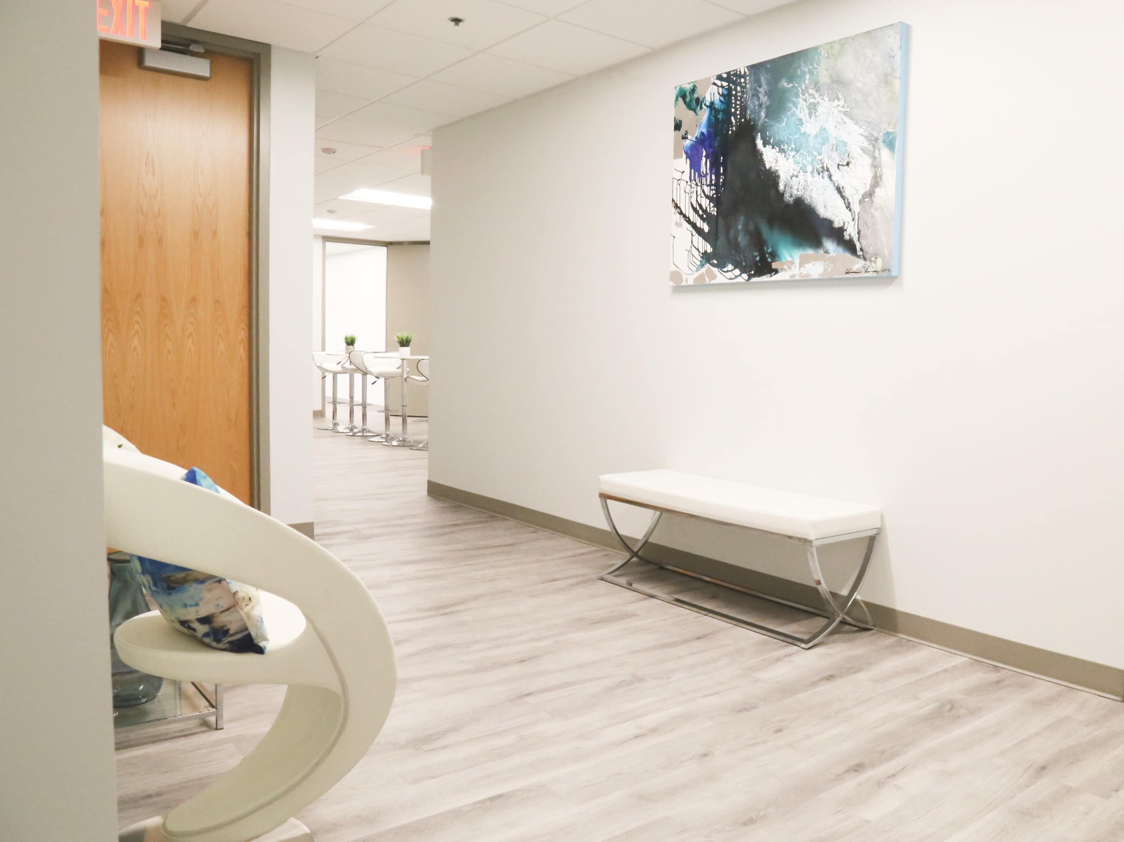 The image shows a modern hallway with light-colored walls, a bench on the floor, and an abstract painting hanging above, leading to a bright area with tables and chairs in the background.