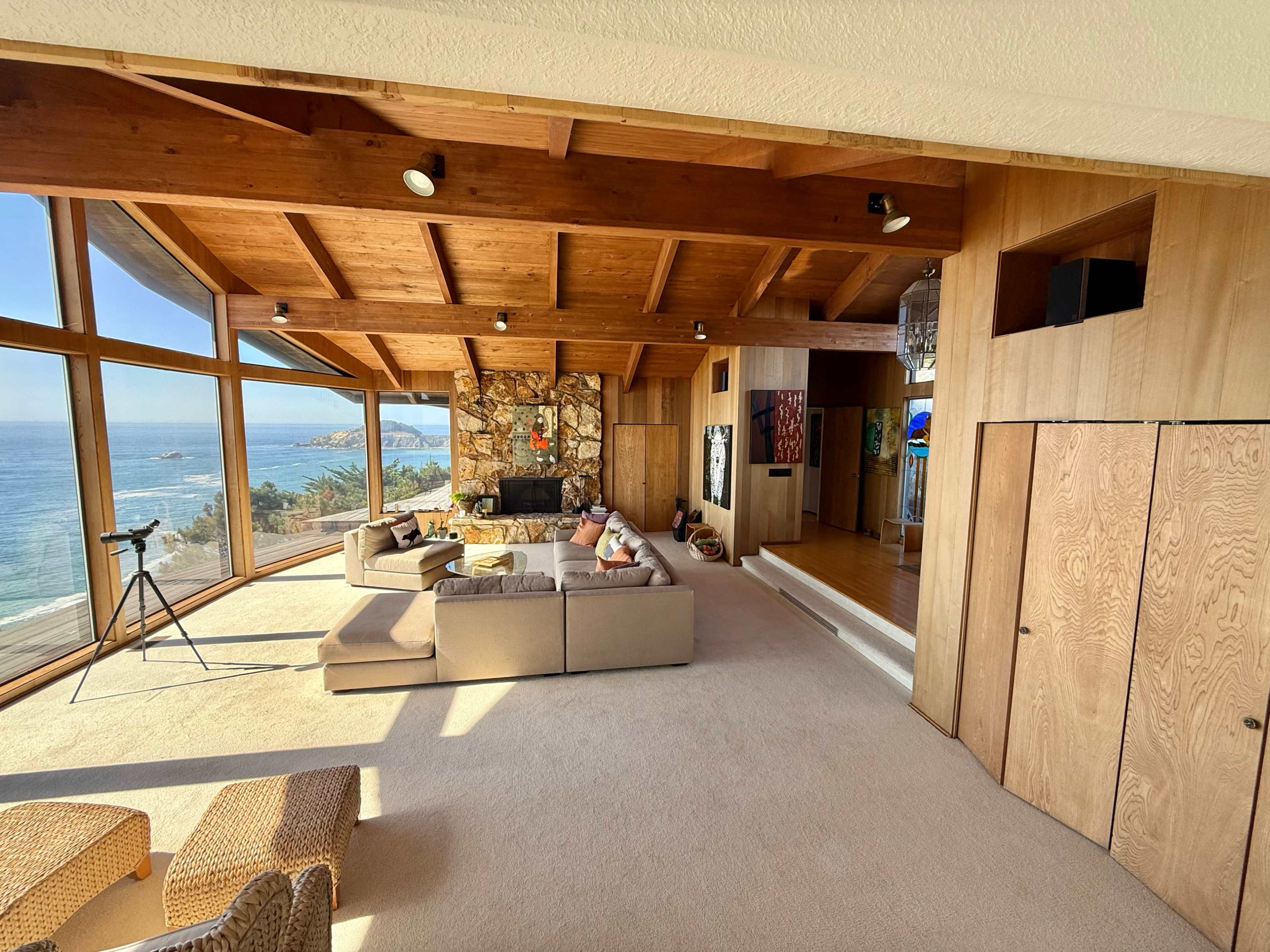 The image shows a spacious living room with a wooden ceiling, large windows overlooking a coastal view, and a stone fireplace as the focal point.