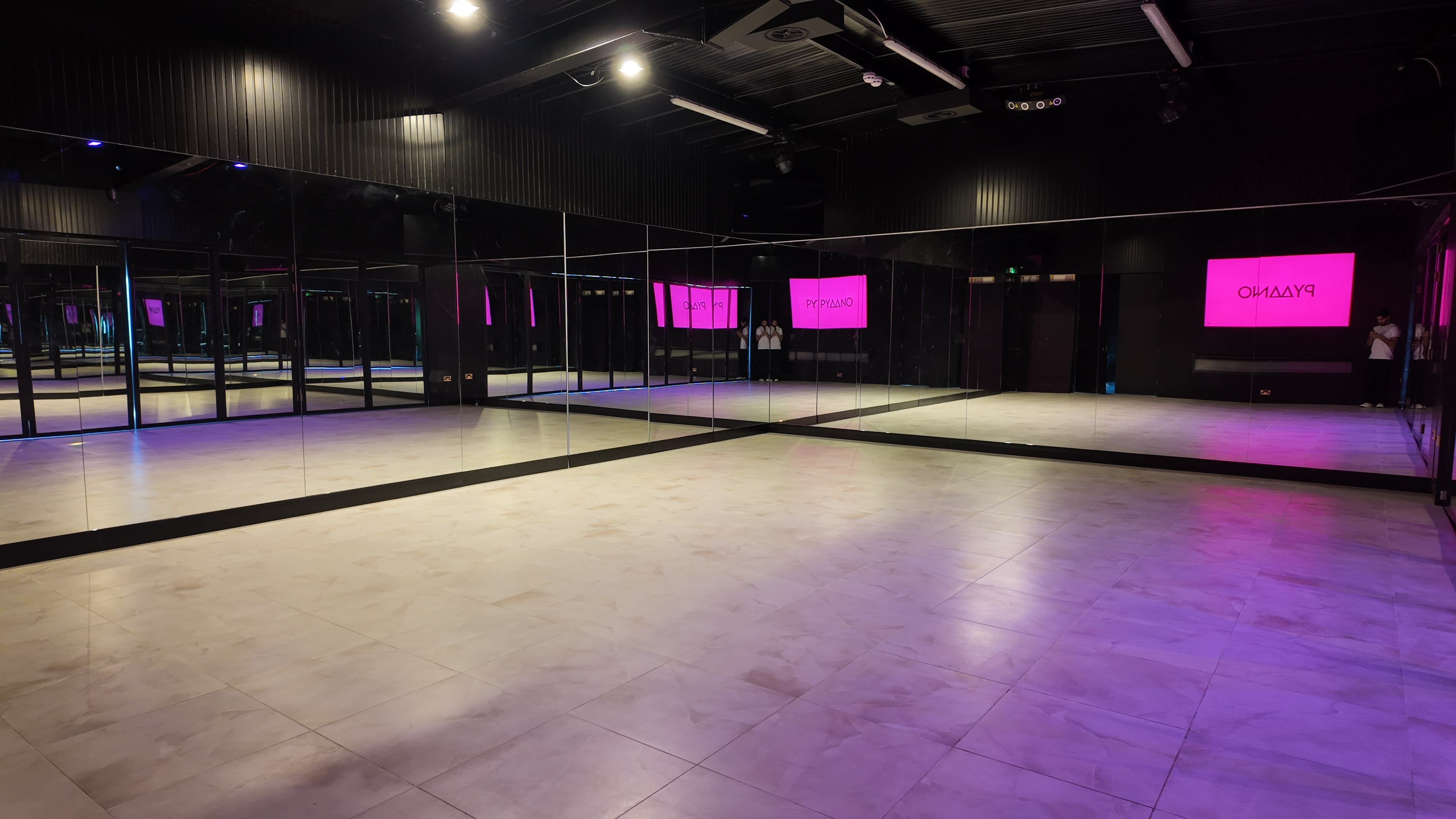 The image shows a spacious dance studio with large mirrors on one wall and neon pink signage in the background.