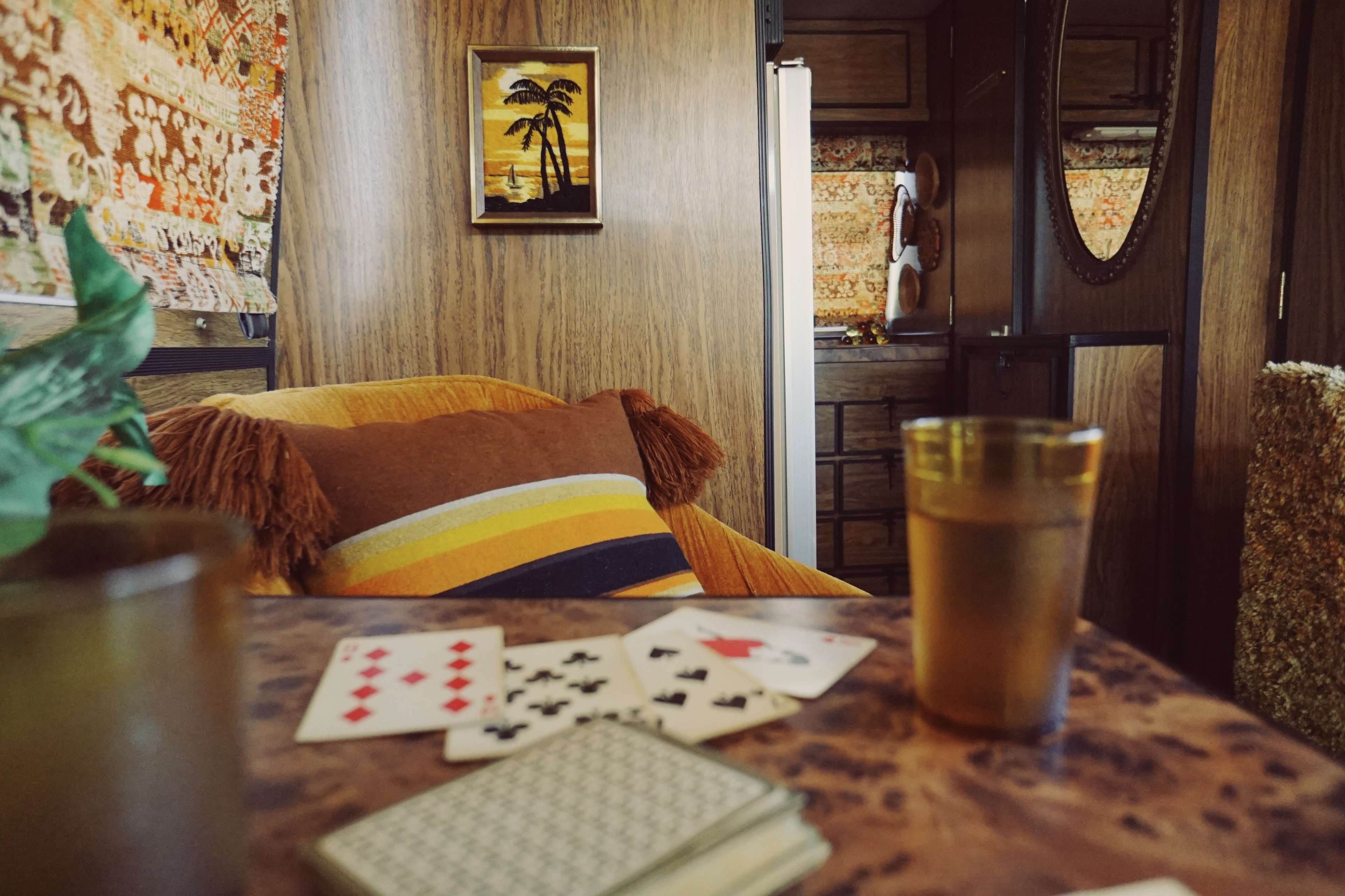 A cozy interior features a wooden table with playing cards and a glass of iced tea, alongside an orange chair and patterned wallpaper.