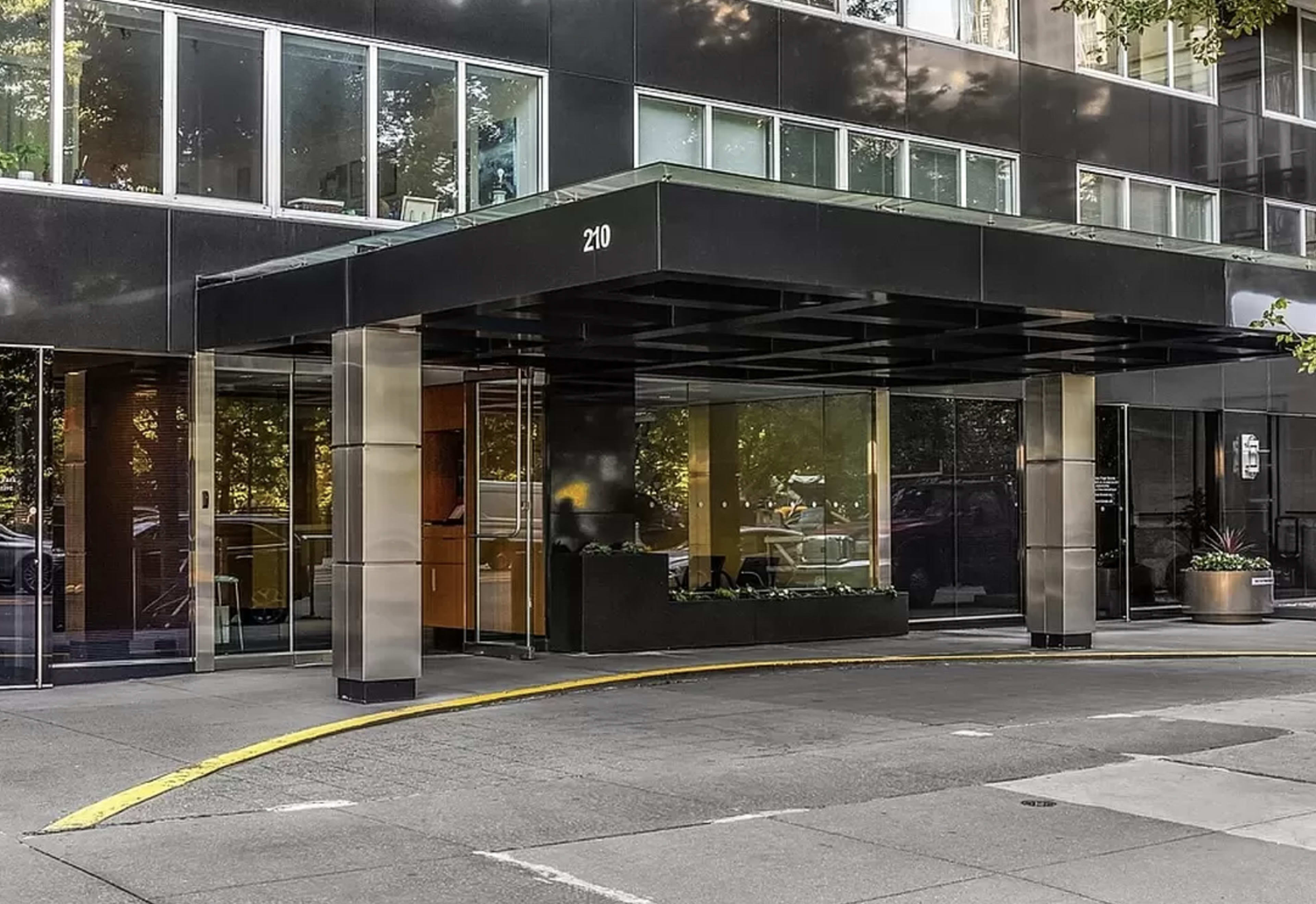 The image shows a modern building entrance with a black canopy and glass walls, featuring large columns and a yellow-curved driveway.