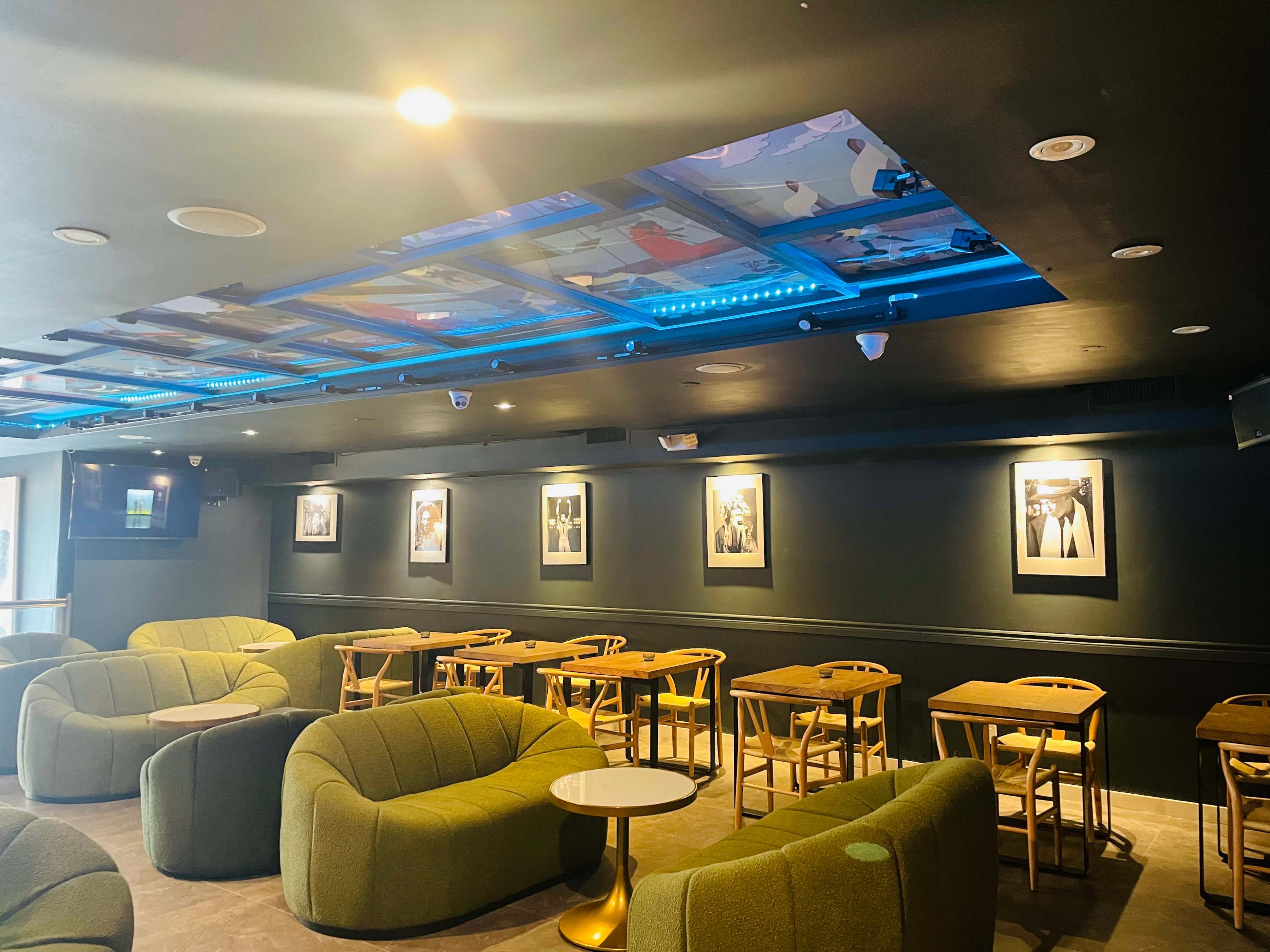 A modern café interior with green upholstered seating, wooden tables, and framed artworks on dark walls, illuminated by blue ceiling lights.