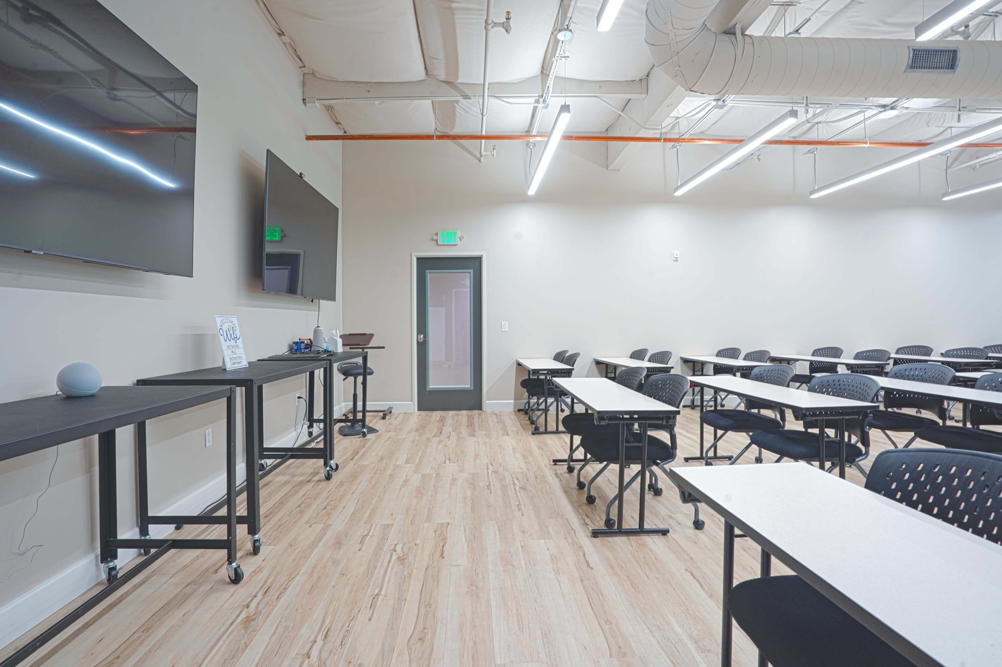A modern classroom features rows of tables and chairs, with large screens mounted on the walls and bright overhead lighting.