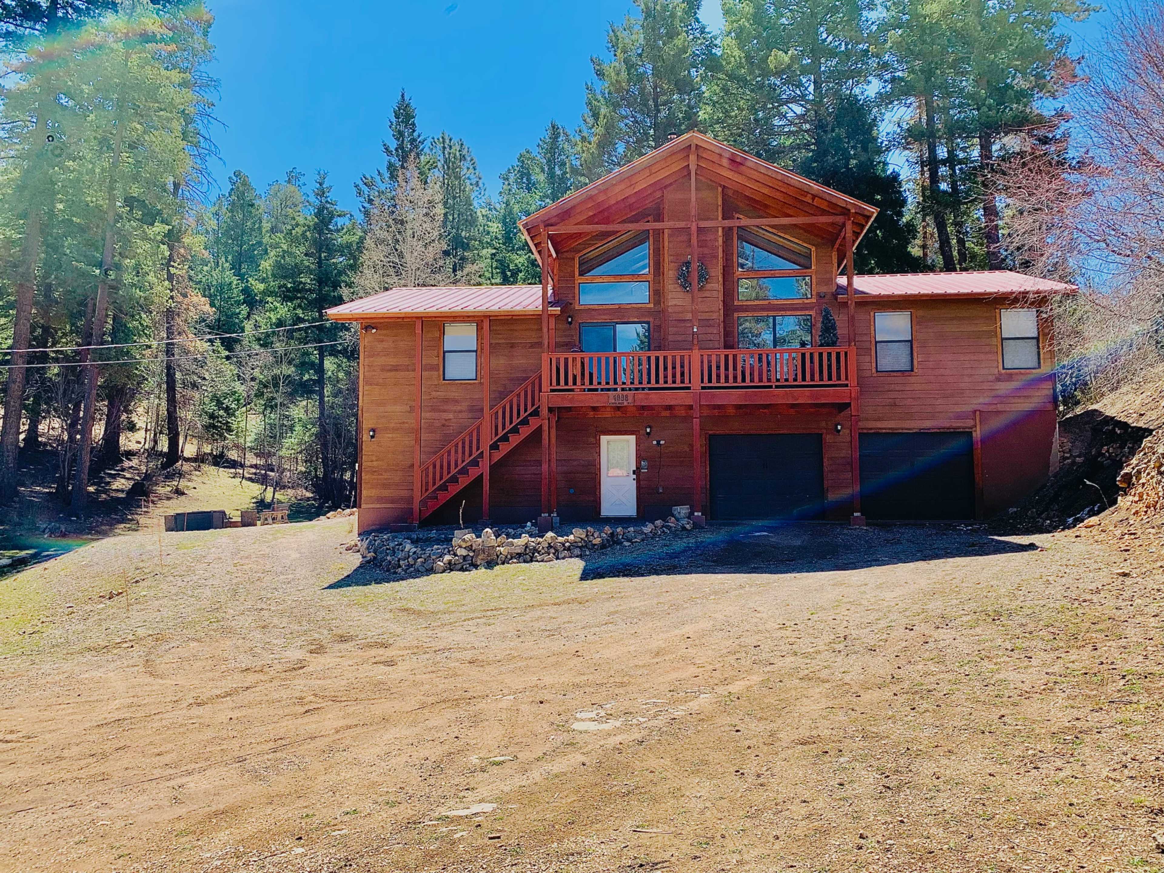 The image shows a two-story wooden house with a red roof surrounded by trees and situated on a gravel driveway.