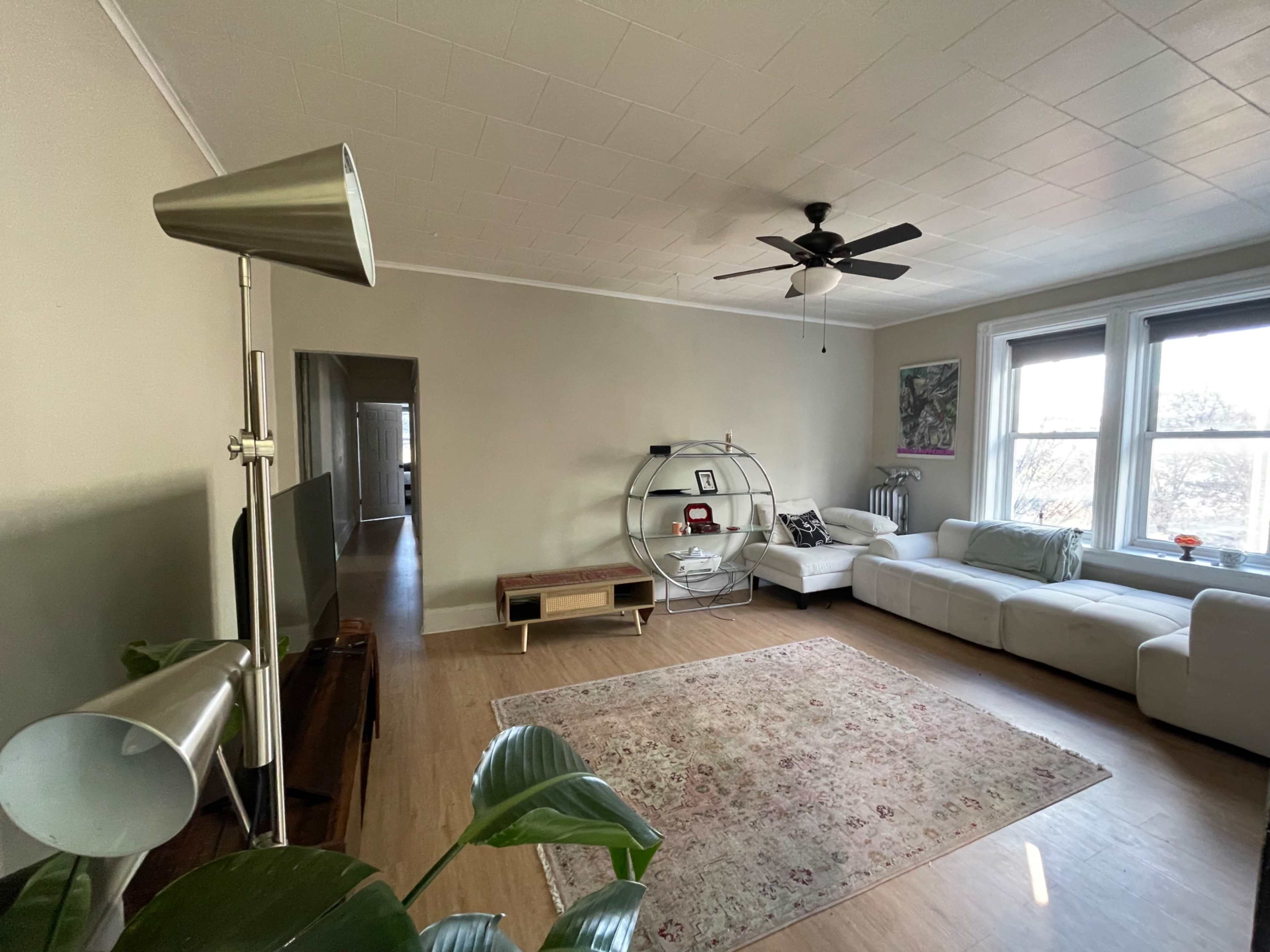 The living room features a white sectional sofa, a round shelving unit, a flat-screen TV on a wooden stand, and a floor lamp, with large windows providing natural light.