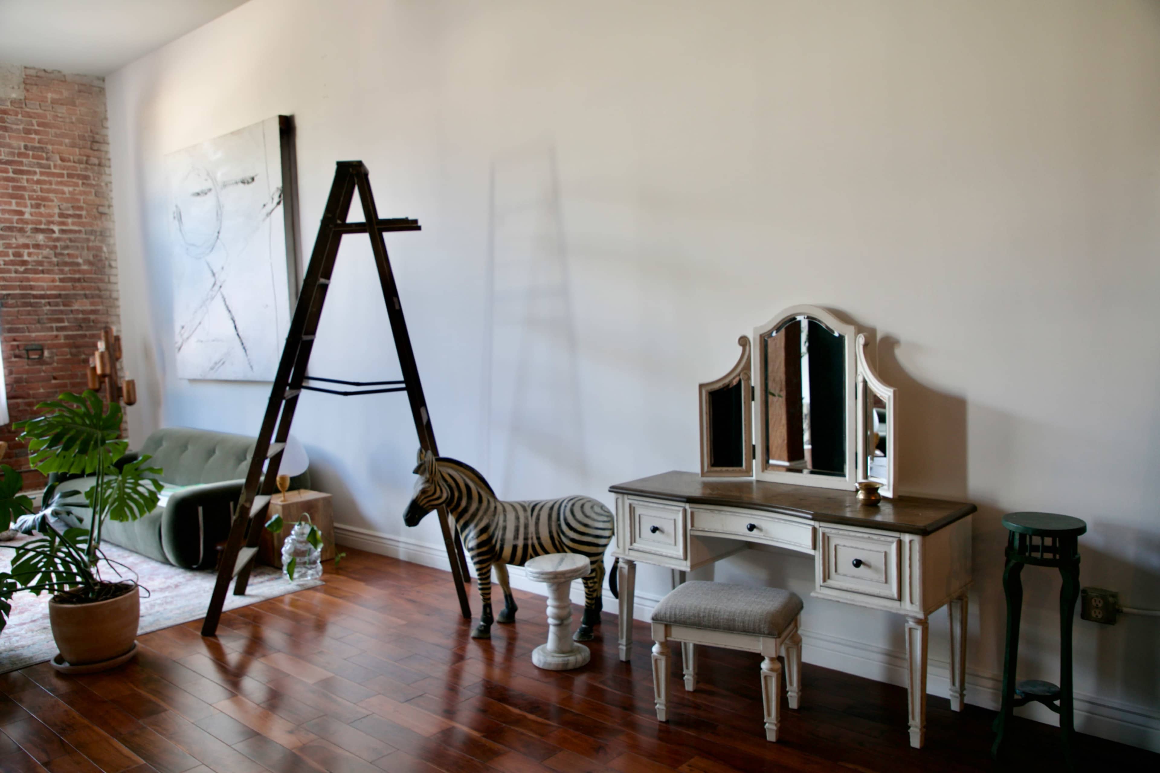 A wooden ladder stands against a wall beside a zebra-striped toy, a vanity table with three mirrors, and a potted plant on a wooden floor.