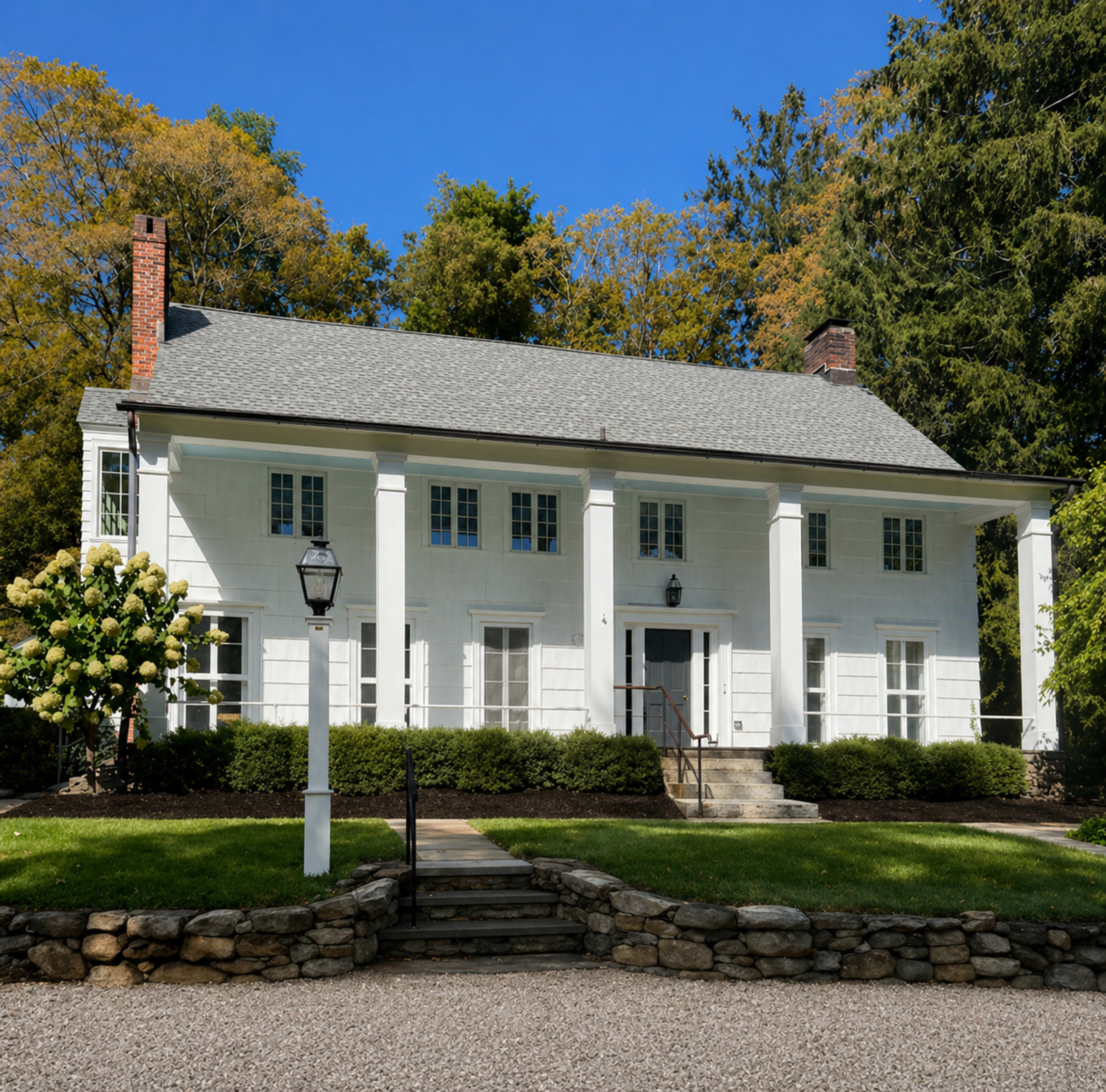 The image shows a two-story white house with a symmetrical facade, featuring a front porch supported by four columns and surrounded by a landscaped yard.