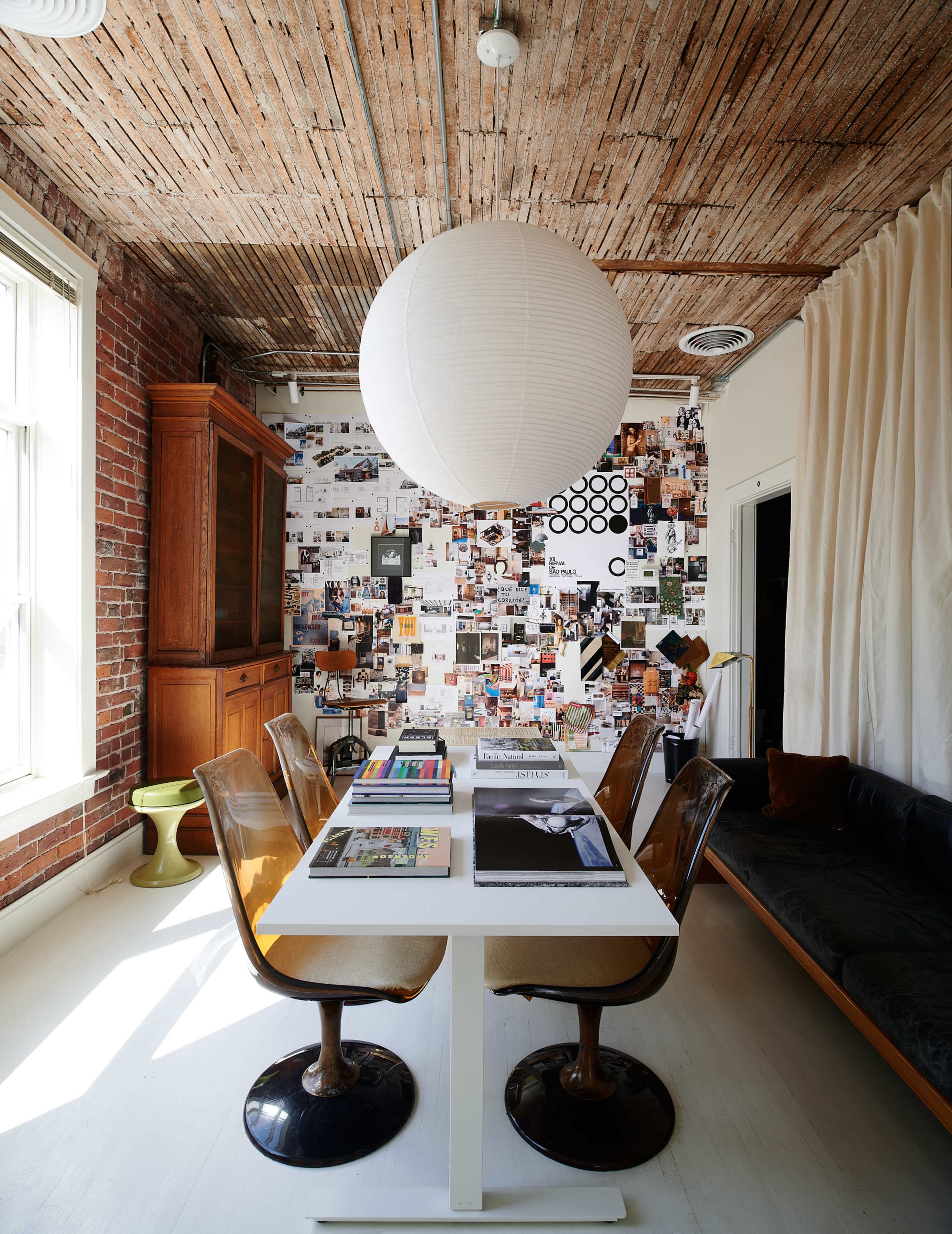A dining area with a central table, surrounded by chairs, a wall covered in photographs and art, and a large paper lantern hanging overhead.