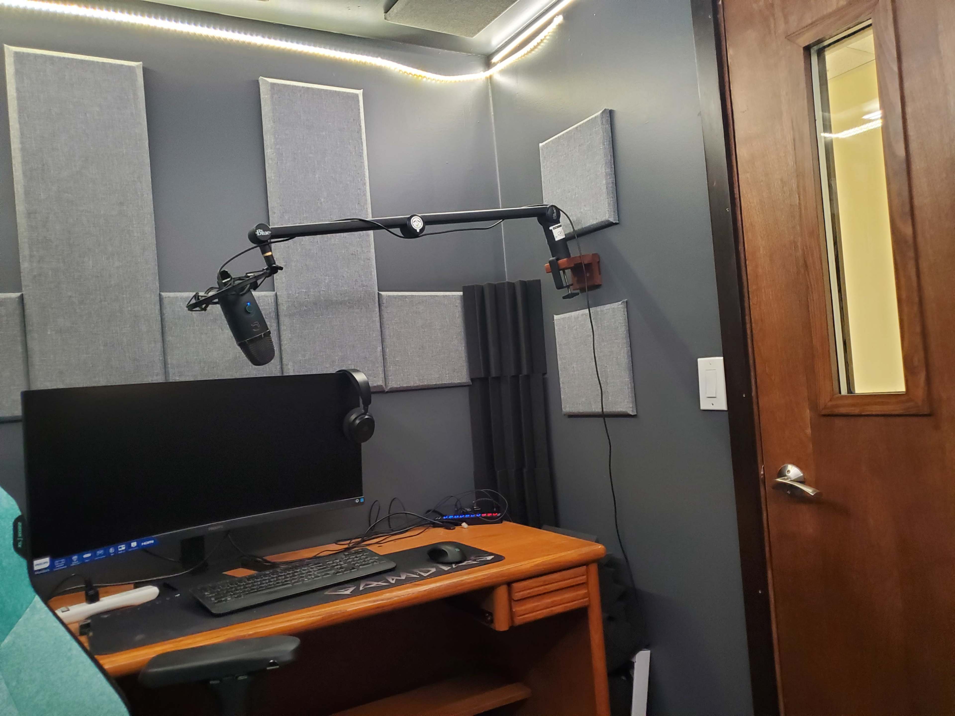 A recording studio setup with a computer monitor, microphone on an adjustable arm, headphones, and soundproofing panels on the walls.