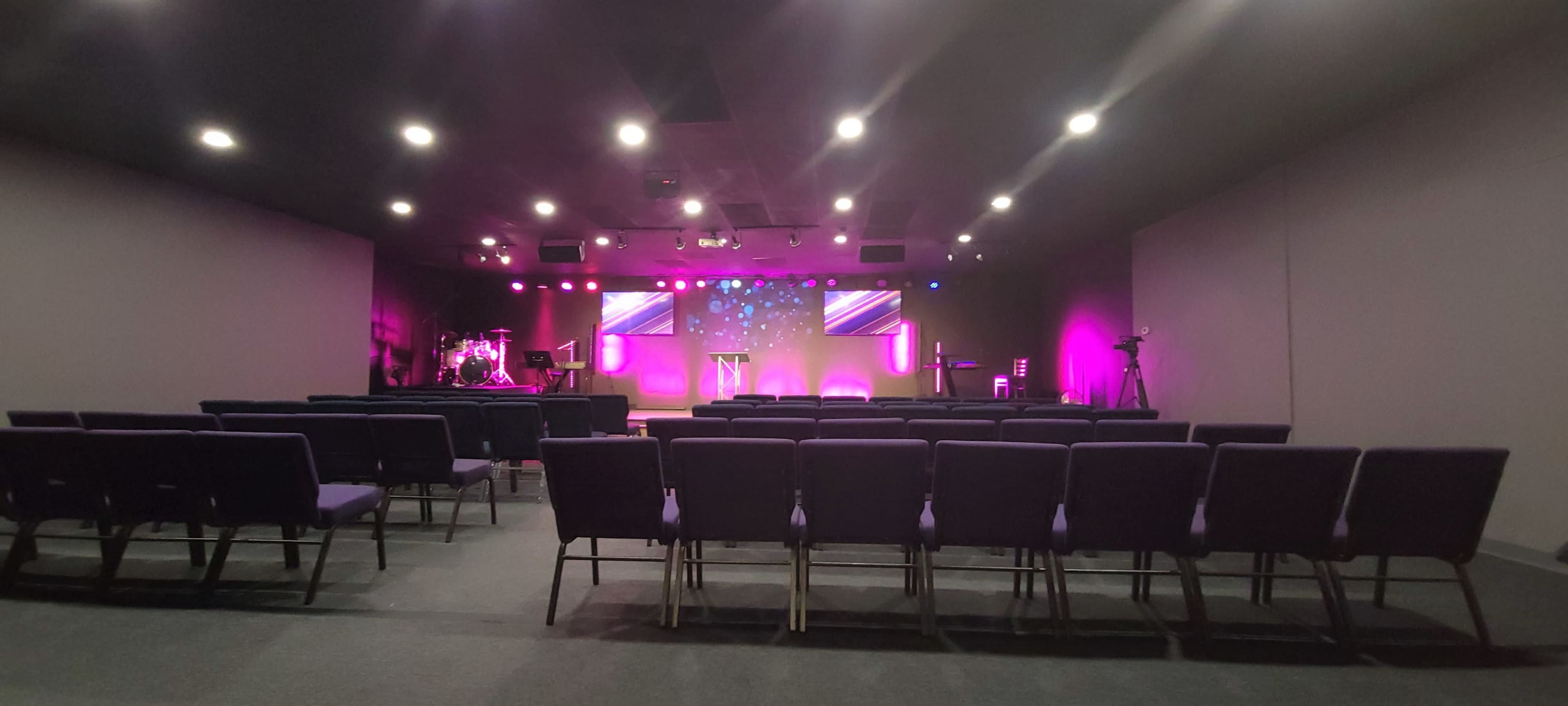 The image shows a modern church auditorium with purple-lit seating and a stage featuring screens and musical instruments in the background.