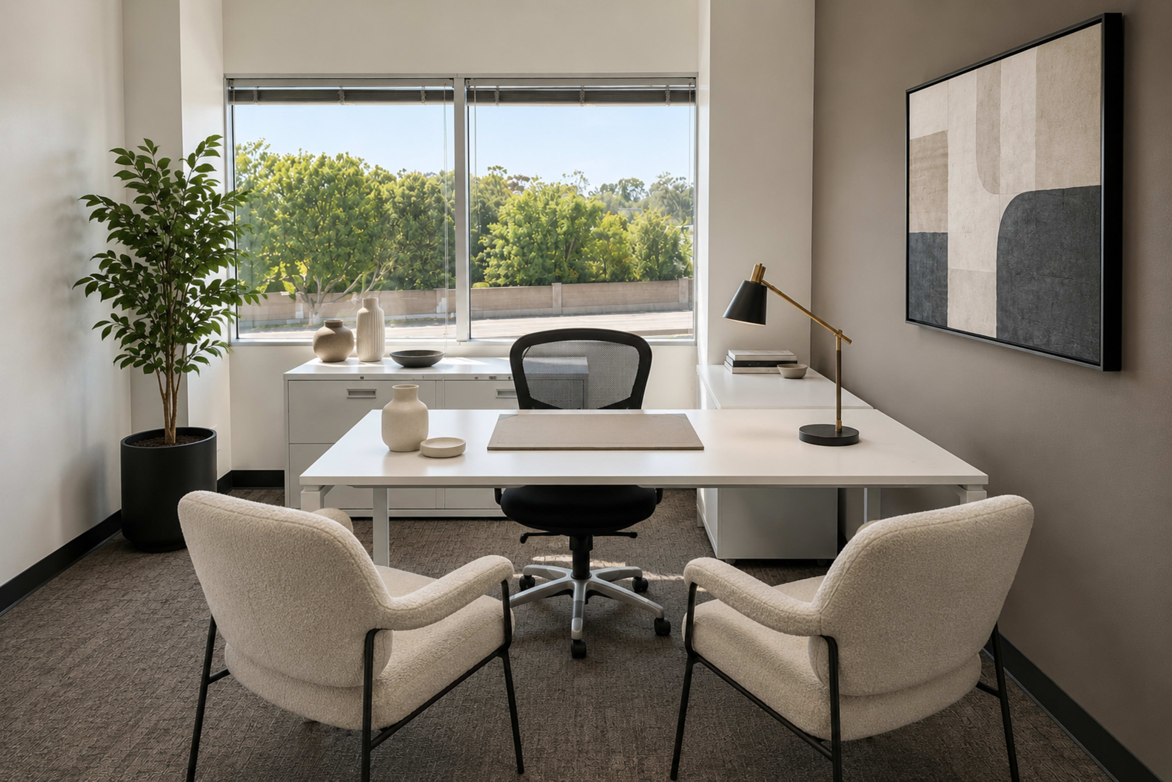 A modern office space features a white desk with a black chair, two light-colored armchairs, a potted plant, and a large window offering a view of greenery outside.