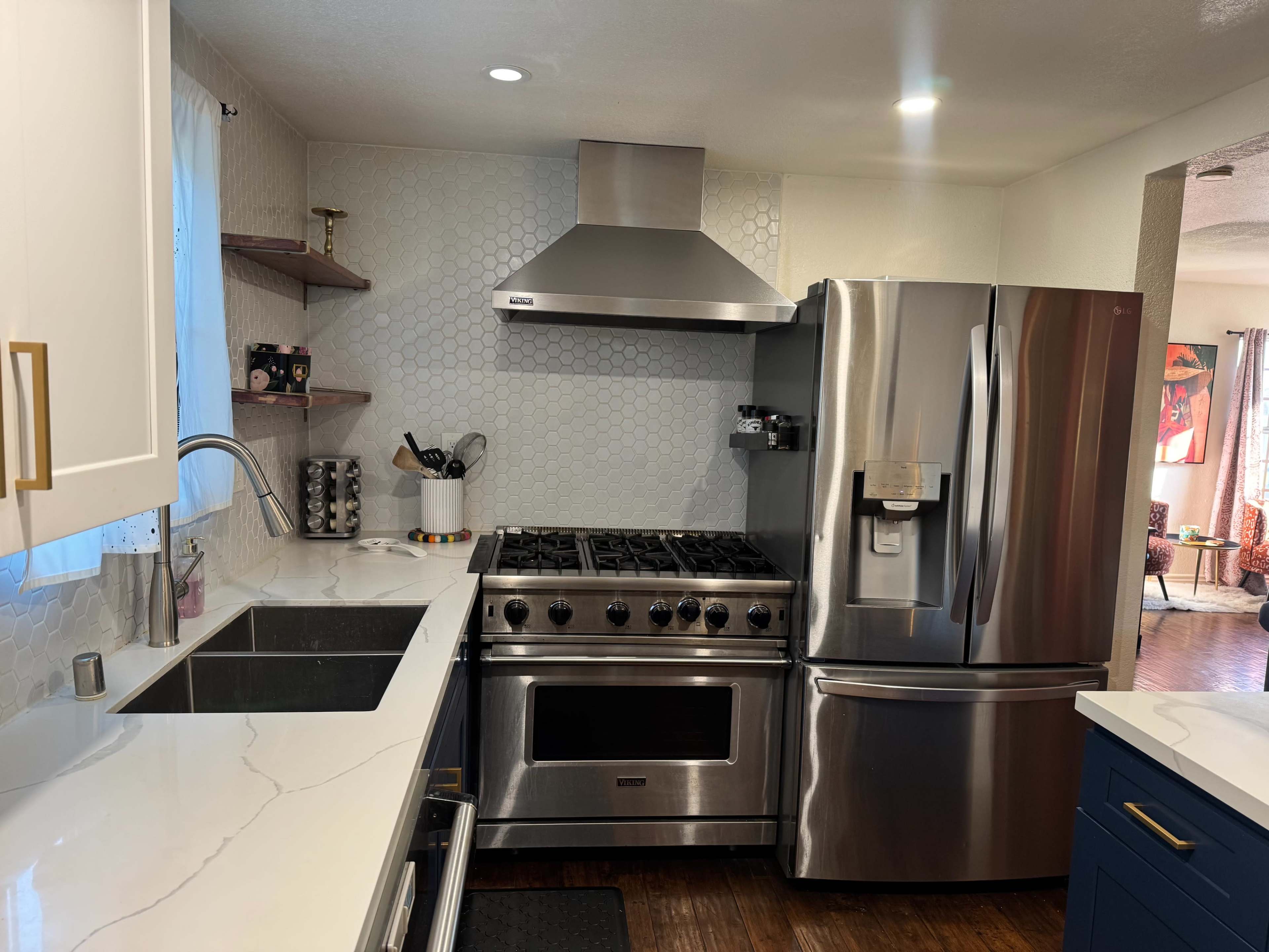 The image shows a modern kitchen featuring a stainless steel gas range, a refrigerator, white cabinets, and a large sink with a marble countertop.