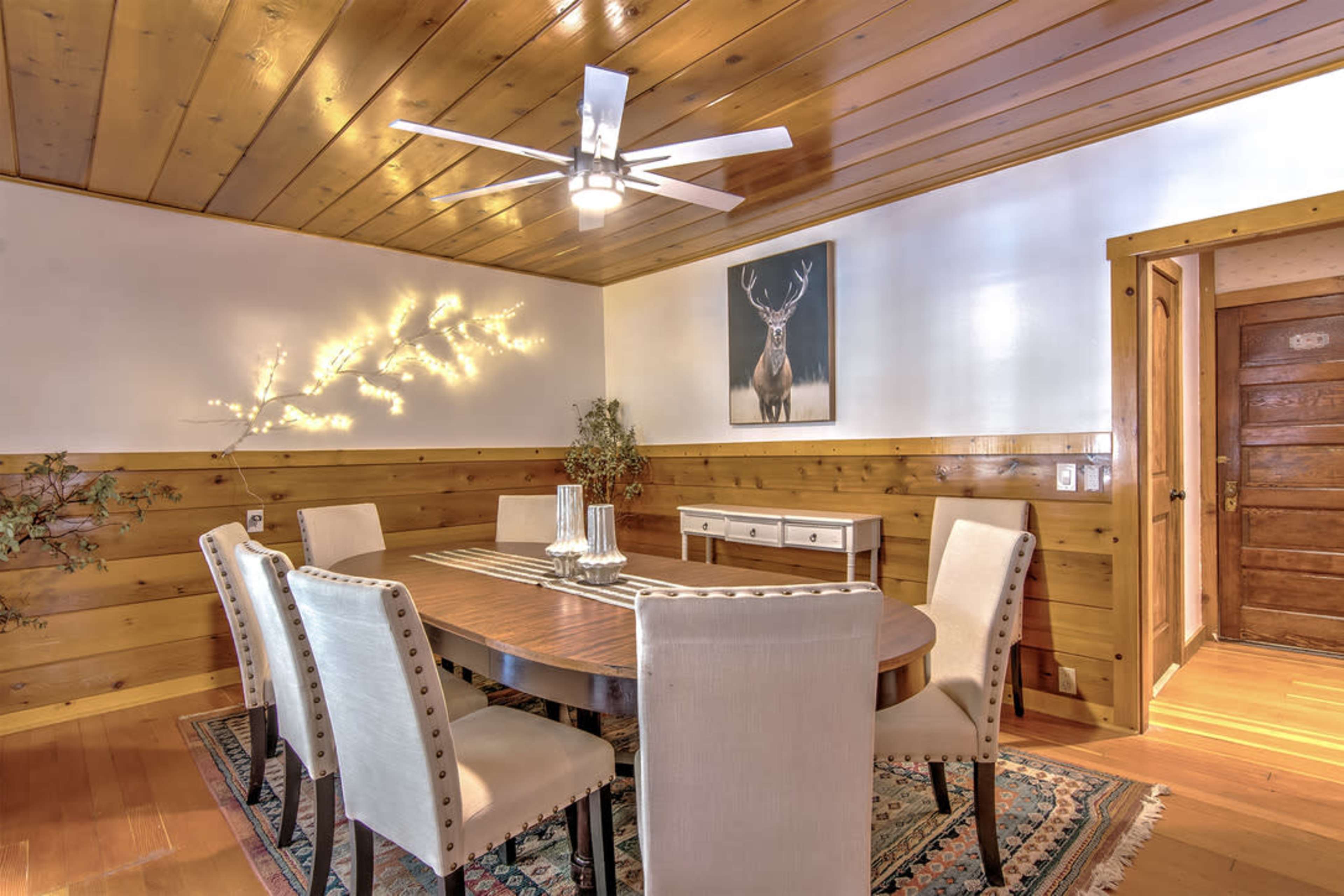 A dining room features a round wooden table surrounded by six upholstered chairs, with a ceiling fan and a wall-mounted deer portrait above a sideboard.
