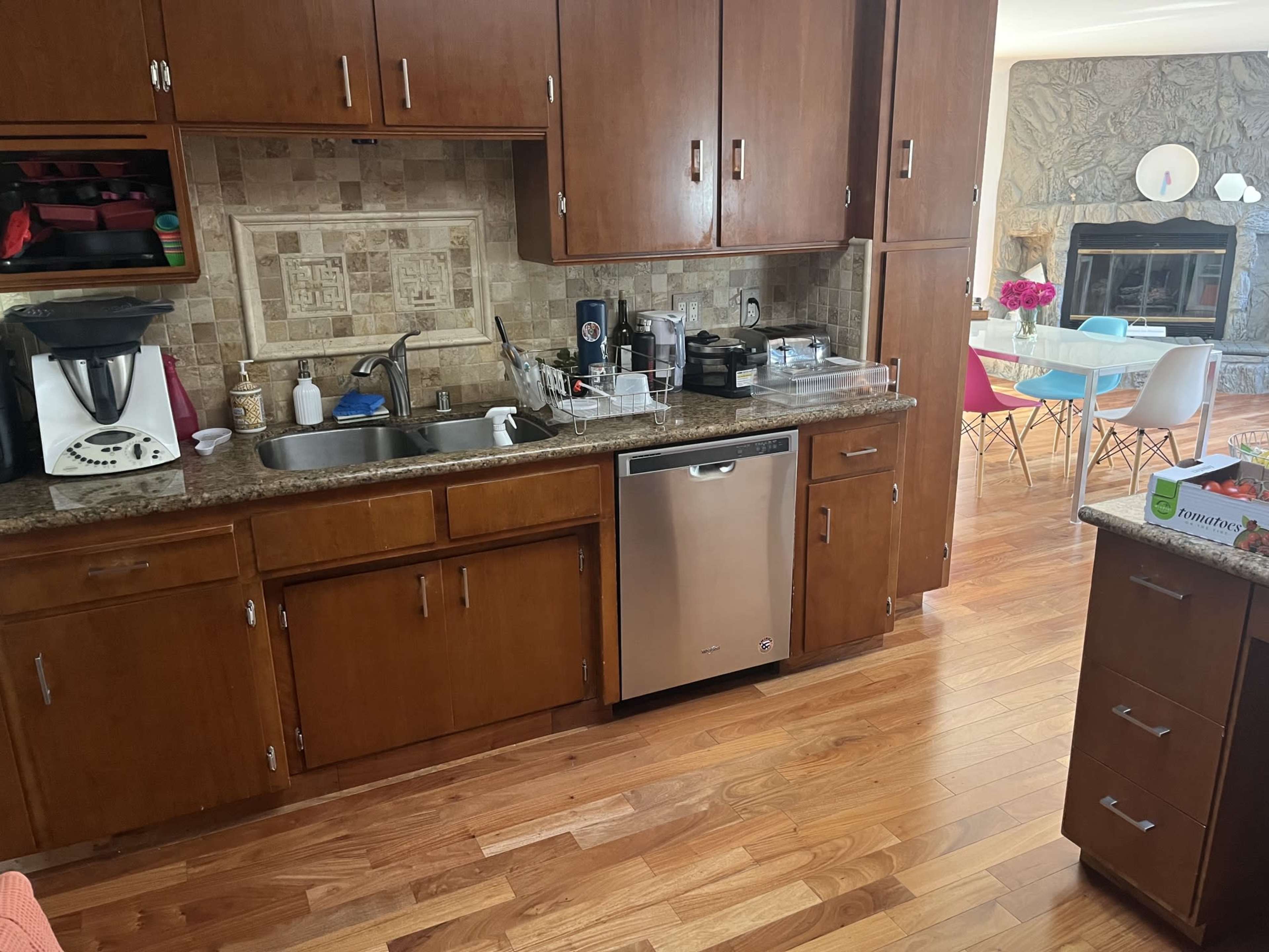 The image shows a kitchen with wooden cabinets, a stainless steel dishwasher, a dual sink, and a countertop with a coffee maker and other kitchen items, while a dining area with colored chairs is visible in the background.